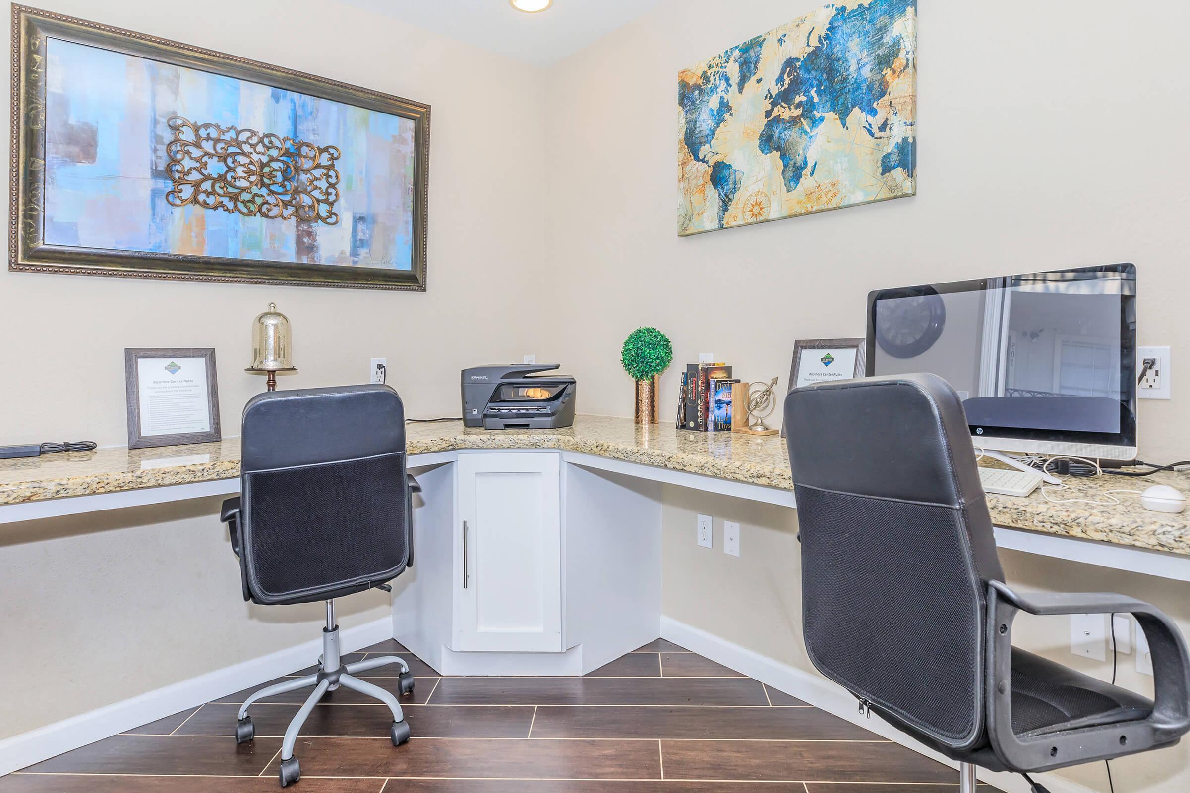 A modern home office setup featuring two black swivel chairs at a long, granite-topped desk. On the desk, there is a computer monitor, a printer, framed documents, a small decorative tree, and a decorative wall art piece depicting a world map. The walls are painted a neutral color, creating a professional ambiance.