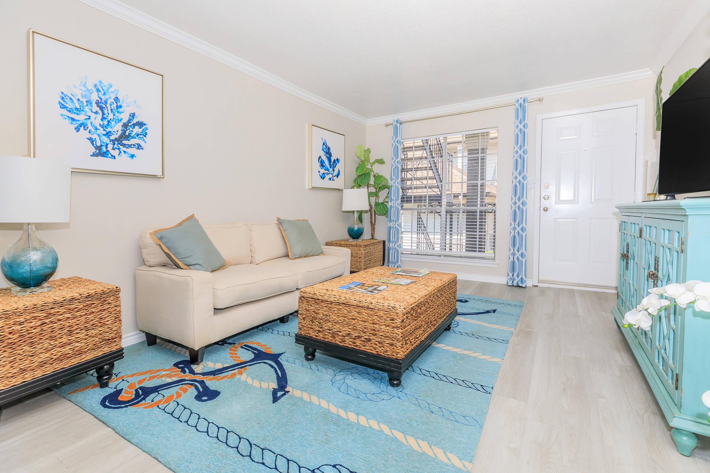 Cozy living room featuring a beige sofa with blue accent pillows, two woven coffee tables, and vibrant coral-themed artwork on the walls. The floor is adorned with a blue area rug, and natural light streams through a window with light curtains. A turquoise cabinet adds a pop of color to the space.