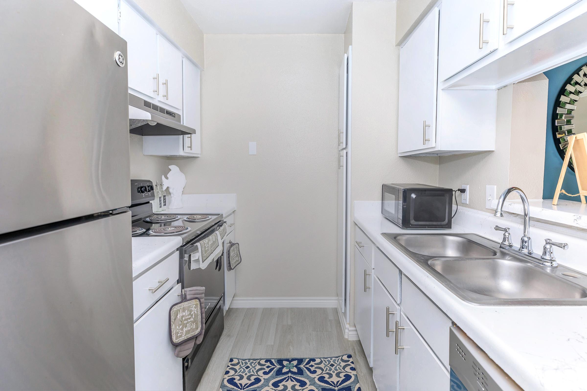 A modern kitchen featuring stainless steel appliances, including a refrigerator and oven, white cabinetry, and a double sink. The space is well-lit with pale walls, gray flooring, and a decorative blue and white rug on the floor. A microwave is placed on the countertop.