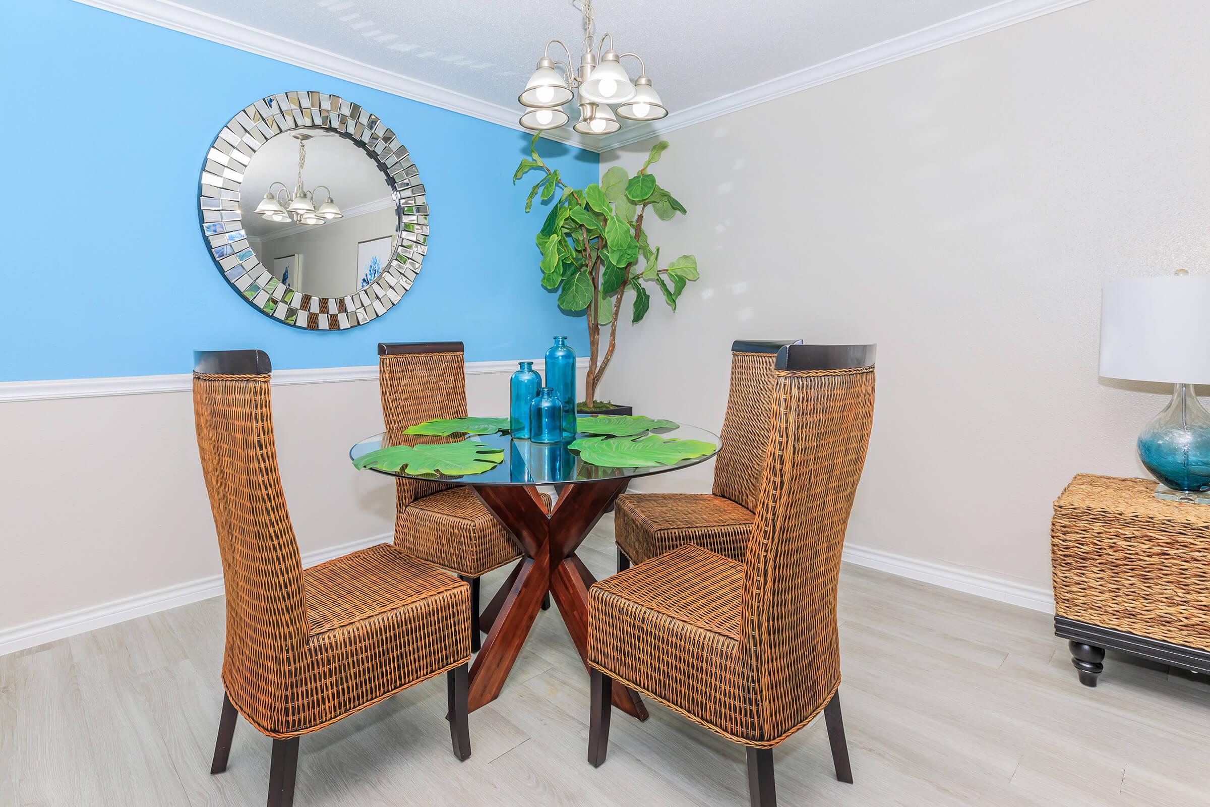 A modern dining area featuring a round glass table with a wooden base, surrounded by four wicker chairs. A decorative mirror hangs on the wall, reflecting light. A potted plant adds a touch of greenery, and blue decorative bottles sit on the table. Soft lighting from a multi-bulb chandelier enhances the inviting atmosphere.