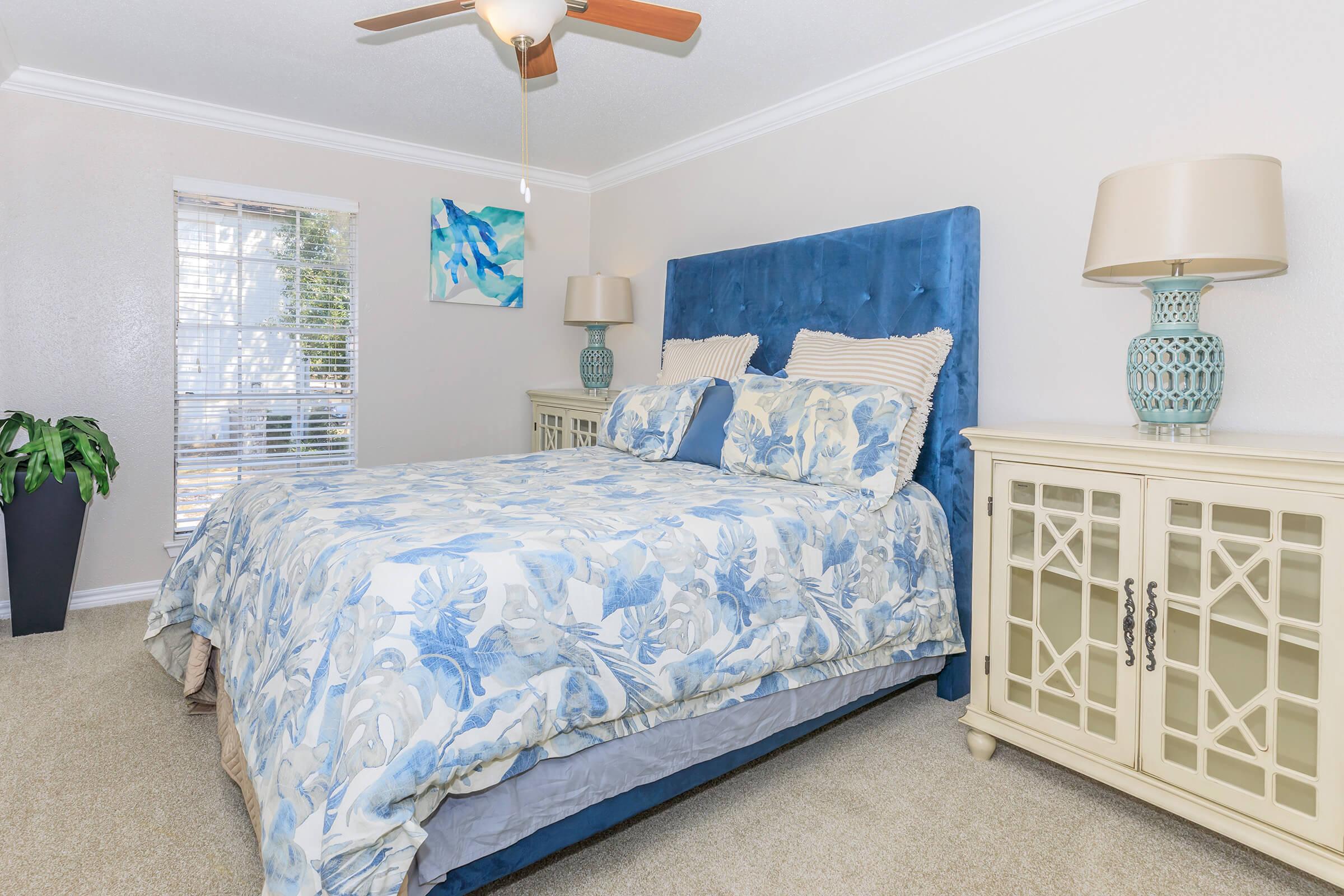 A cozy bedroom featuring a blue floral bedspread on a bed with a tufted blue headboard. Light-colored bedside tables with lamps are on either side, and a decorative piece is on the wall above the bed. A window with white blinds lets in natural light, and a potted plant adds a touch of greenery.
