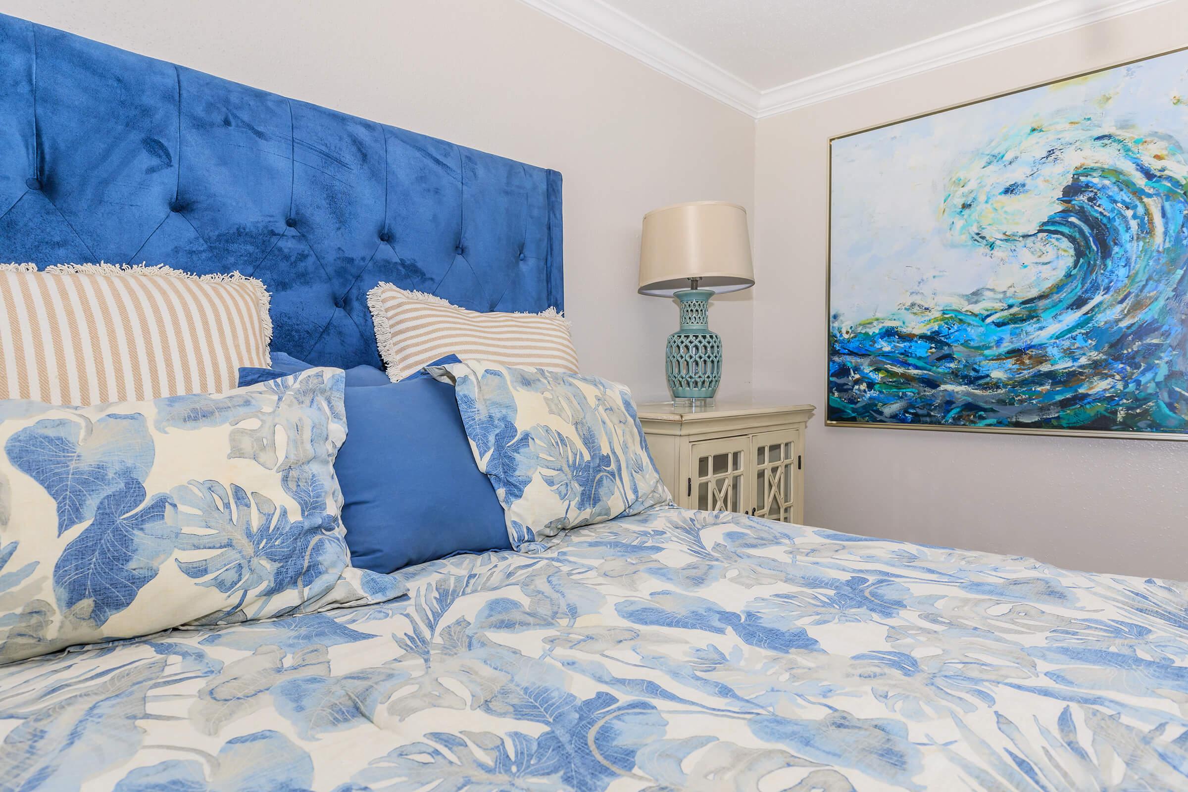 A cozy bedroom featuring a blue tufted headboard, a bed adorned with floral-patterned pillows and bedding, a decorative lamp on a nightstand, and an abstract painting of ocean waves on the wall. The overall color palette includes shades of blue and cream, creating a serene atmosphere.