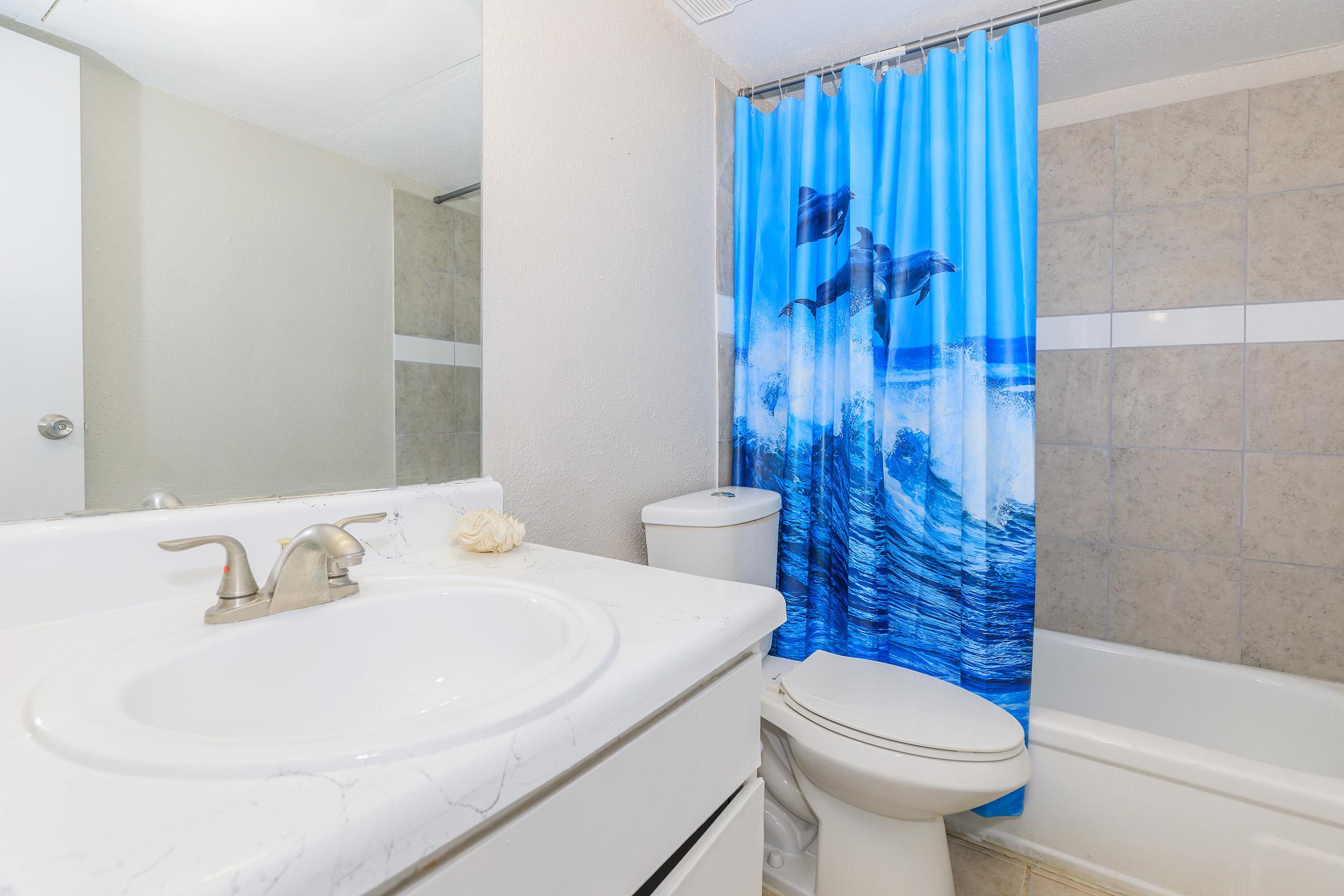 A bathroom featuring a white sink and countertop, a toilet, and a bathtub. The bathtub is adorned with a blue shower curtain displaying dolphins jumping through ocean waves. Natural light reflects off the surfaces, enhancing the clean and bright atmosphere.