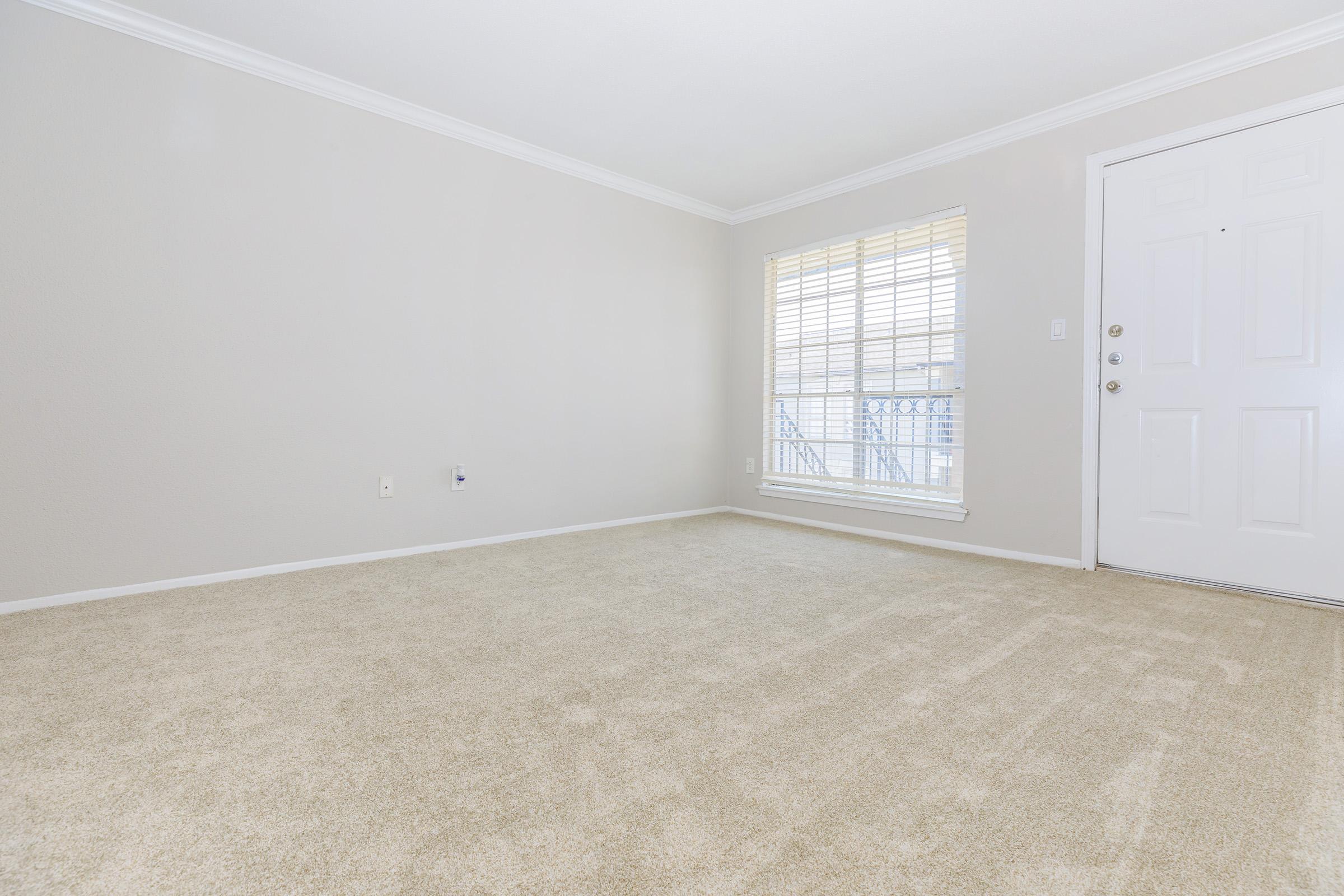 A spacious, empty room with beige carpet and light gray walls. There's a large window with white blinds on the left, allowing natural light in. A white door is visible on the right, leading to the exterior. The room feels bright and welcoming, suitable for various furniture arrangements.