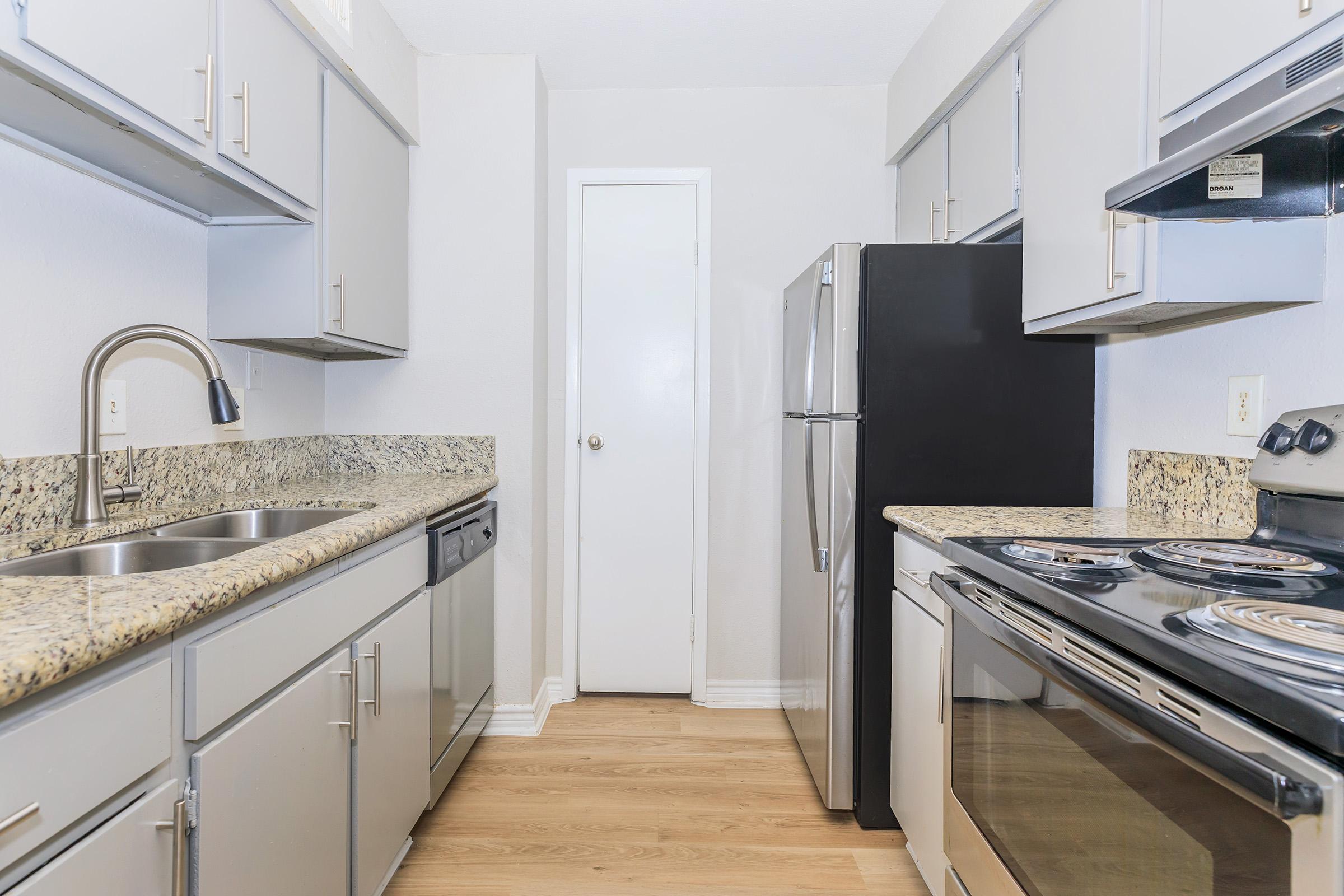 Modern kitchen featuring light gray cabinets, granite countertops, stainless steel appliances including a refrigerator and stove, and a double sink. The walls are light-colored, and there is a door leading to another room. The floor is wooden, creating a warm atmosphere.