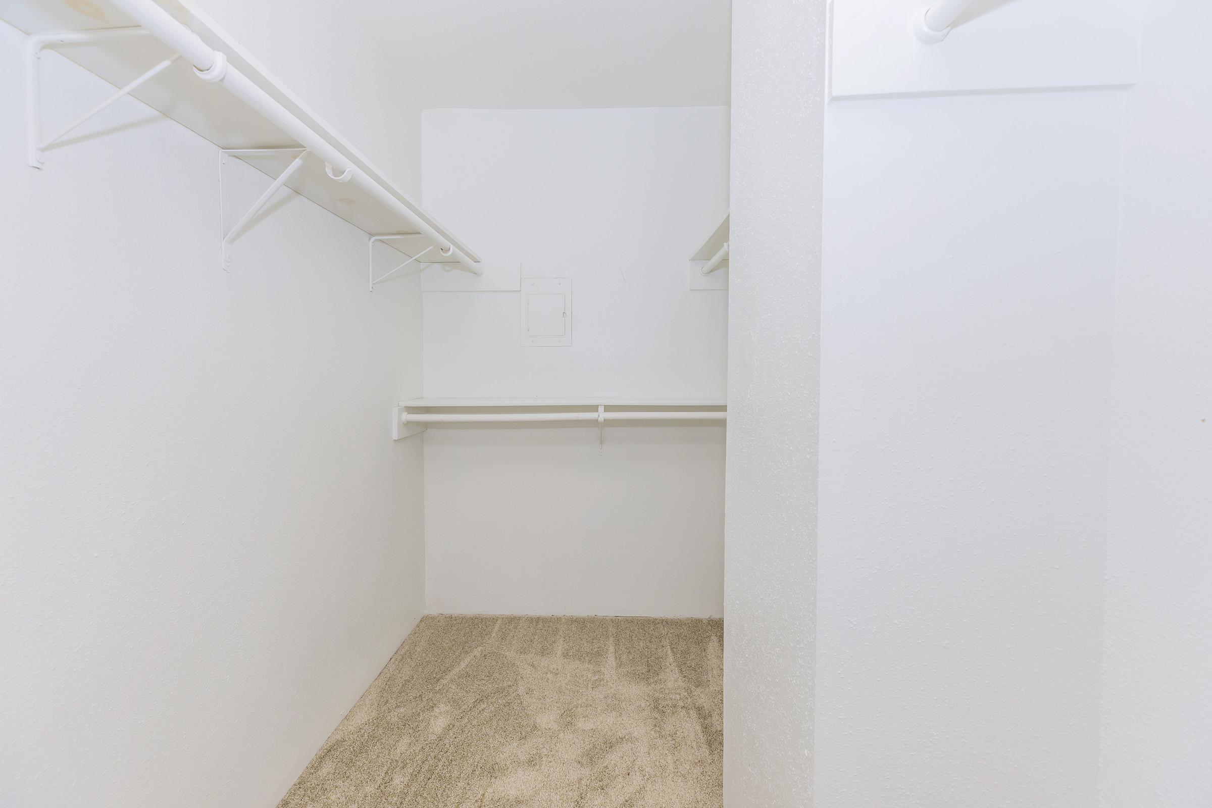 An empty walk-in closet featuring white walls, light carpet flooring, and several hanging rods for storage. The space is well-lit and minimalistic, providing a clean and organized area for clothes and accessories.
