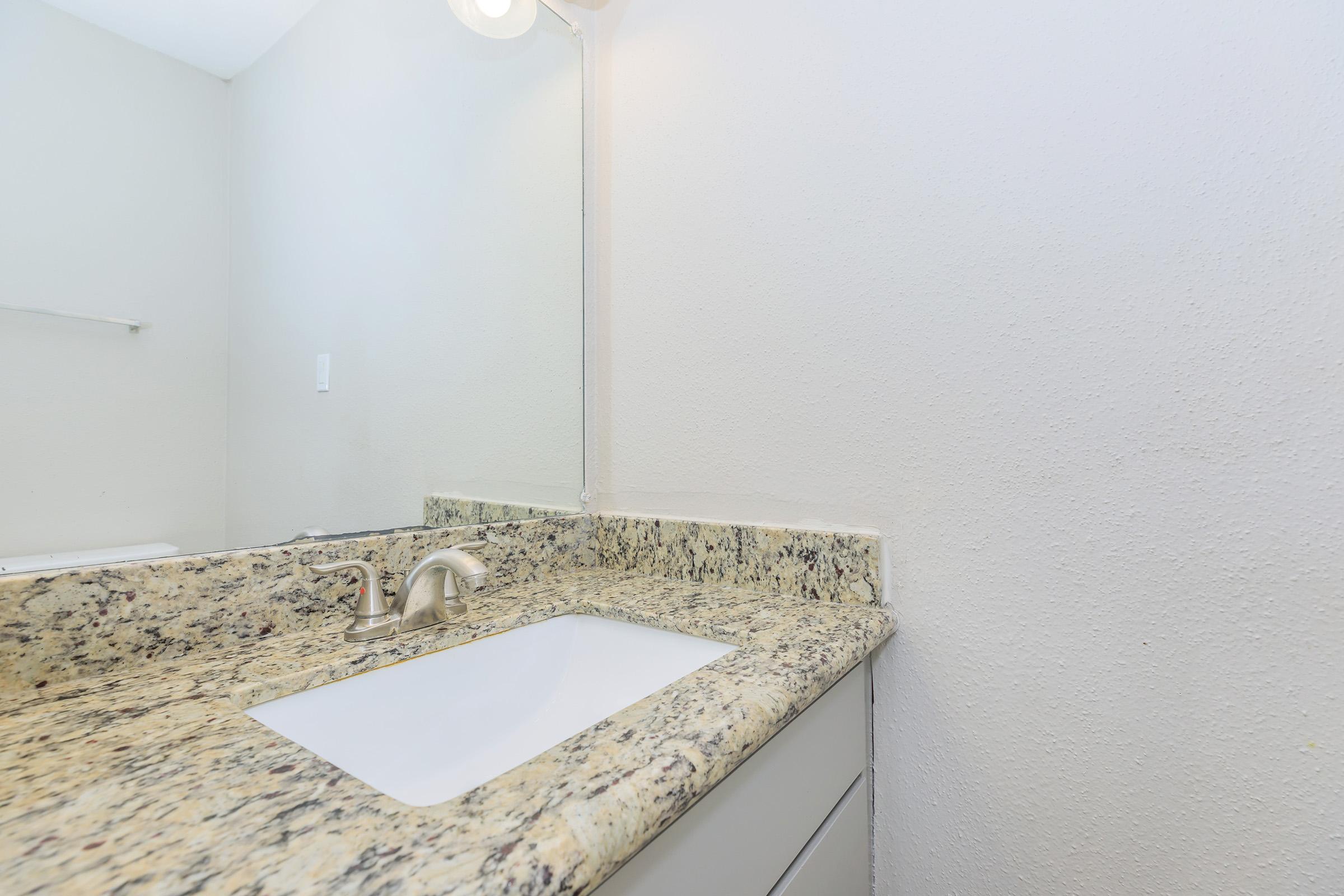 A bathroom countertop featuring a white sink surrounded by a granite surface with speckles. A wall-mounted light fixture is visible above the mirror, and the wall is painted a light color. The space appears clean and modern, with a minimalist design.