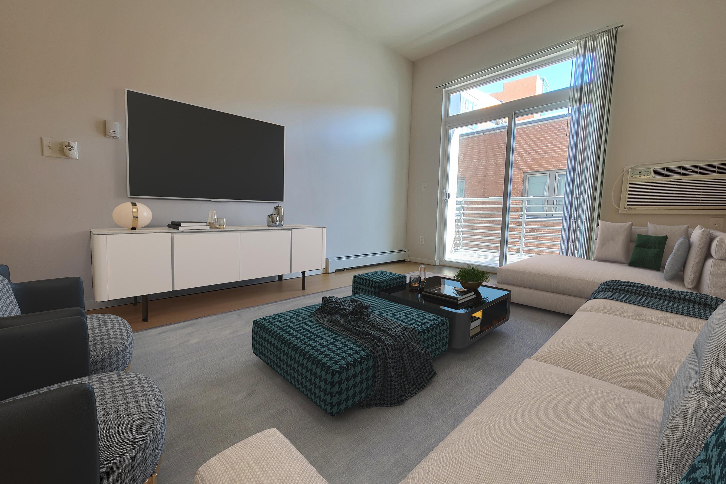 A modern living room featuring a large flat-screen TV mounted on the wall, a white media console, and a light-colored sectional sofa. There are dark blue accent chairs, a coffee table with green and black geometric patterns, and large windows allowing natural light to illuminate the space.