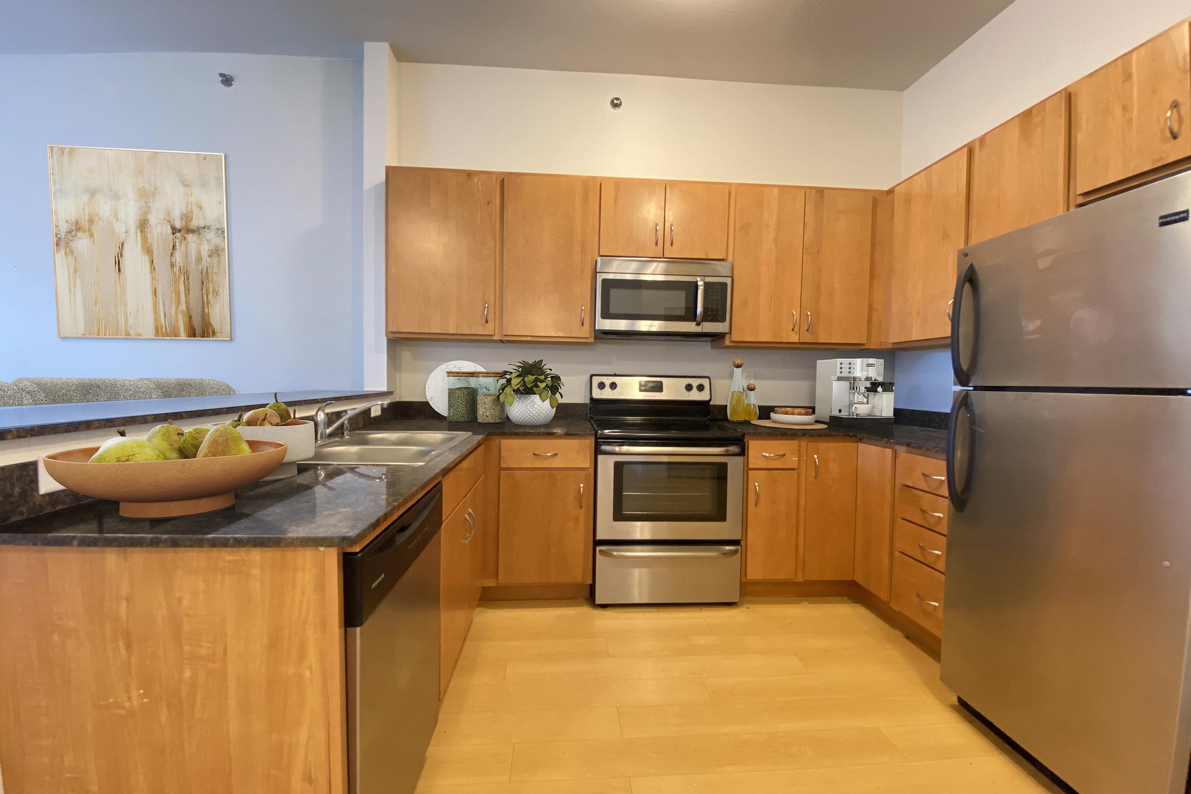 Modern kitchen featuring wooden cabinetry, stainless steel appliances, a granite countertop, and an island with a bowl of pears. The space is well-lit, with light wood flooring and a piece of abstract art on the wall, creating a warm and inviting atmosphere.