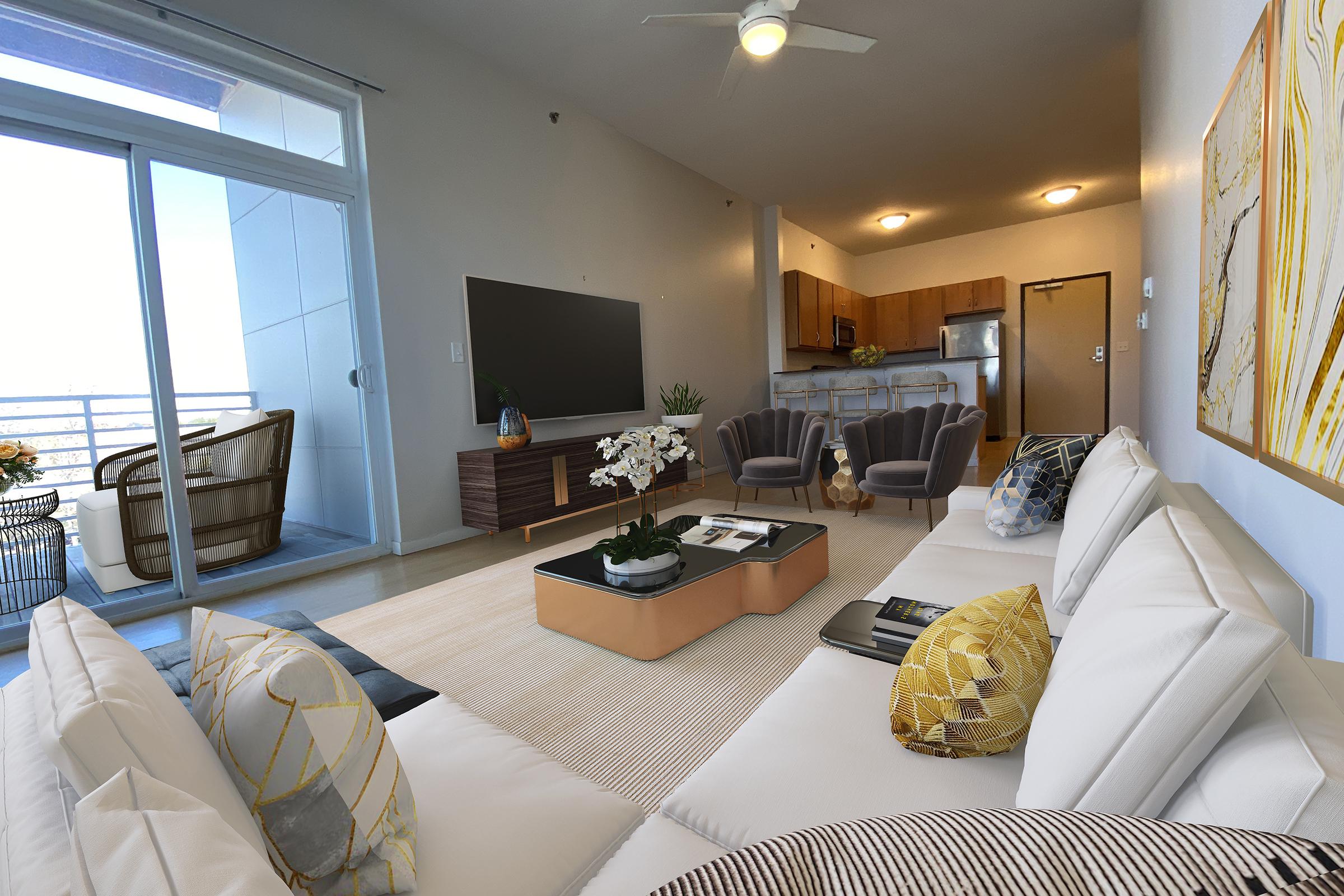 A stylish living room featuring a modern white sofa with decorative pillows, a coffee table with magazines, and a large flat-screen TV. In the background, a kitchen area with wooden cabinetry is visible. The space opens to a balcony with seating, and decorative artwork adorns the walls, enhancing the contemporary aesthetic.