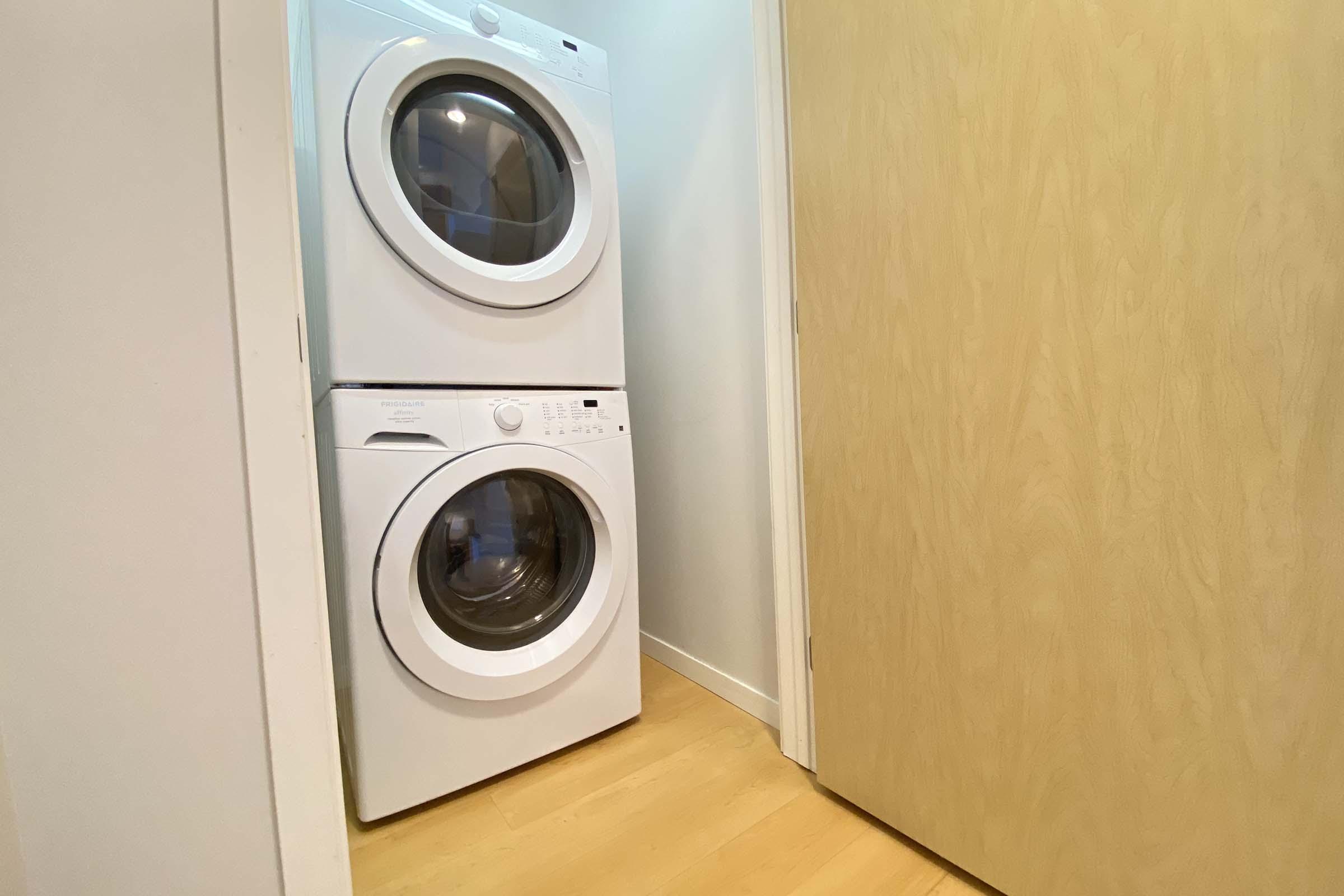 A stackable washer and dryer in a laundry space, with a light-colored wood door partially visible. The interior has neutral walls and wooden flooring, creating a modern and clean look.