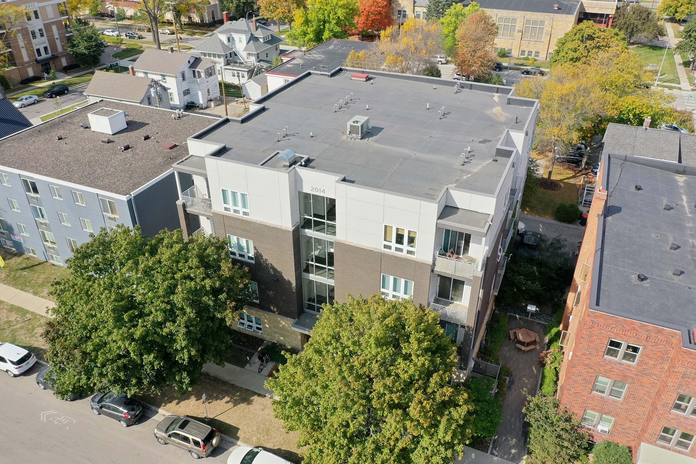 Aerial view of a modern multi-story apartment building surrounded by trees and parked cars. The building features a mix of brick and metal siding, with balconies visible on the upper floors. Nearby residential homes and colorful autumn foliage can be seen in the background.