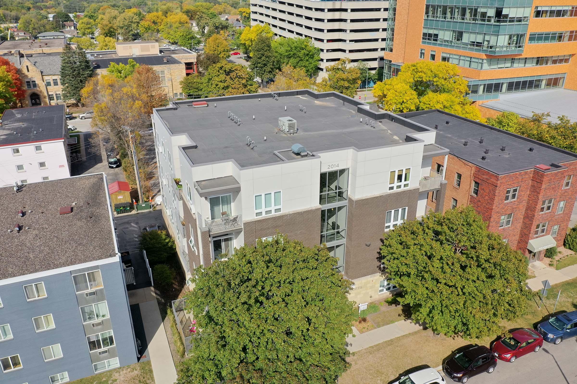 Aerial view of a modern multi-story residential building surrounded by trees and parked cars. The building features a mix of brick and contemporary design elements, with neighboring structures visible in the background. The scene captures a mix of urban and natural elements in an inviting neighborhood.