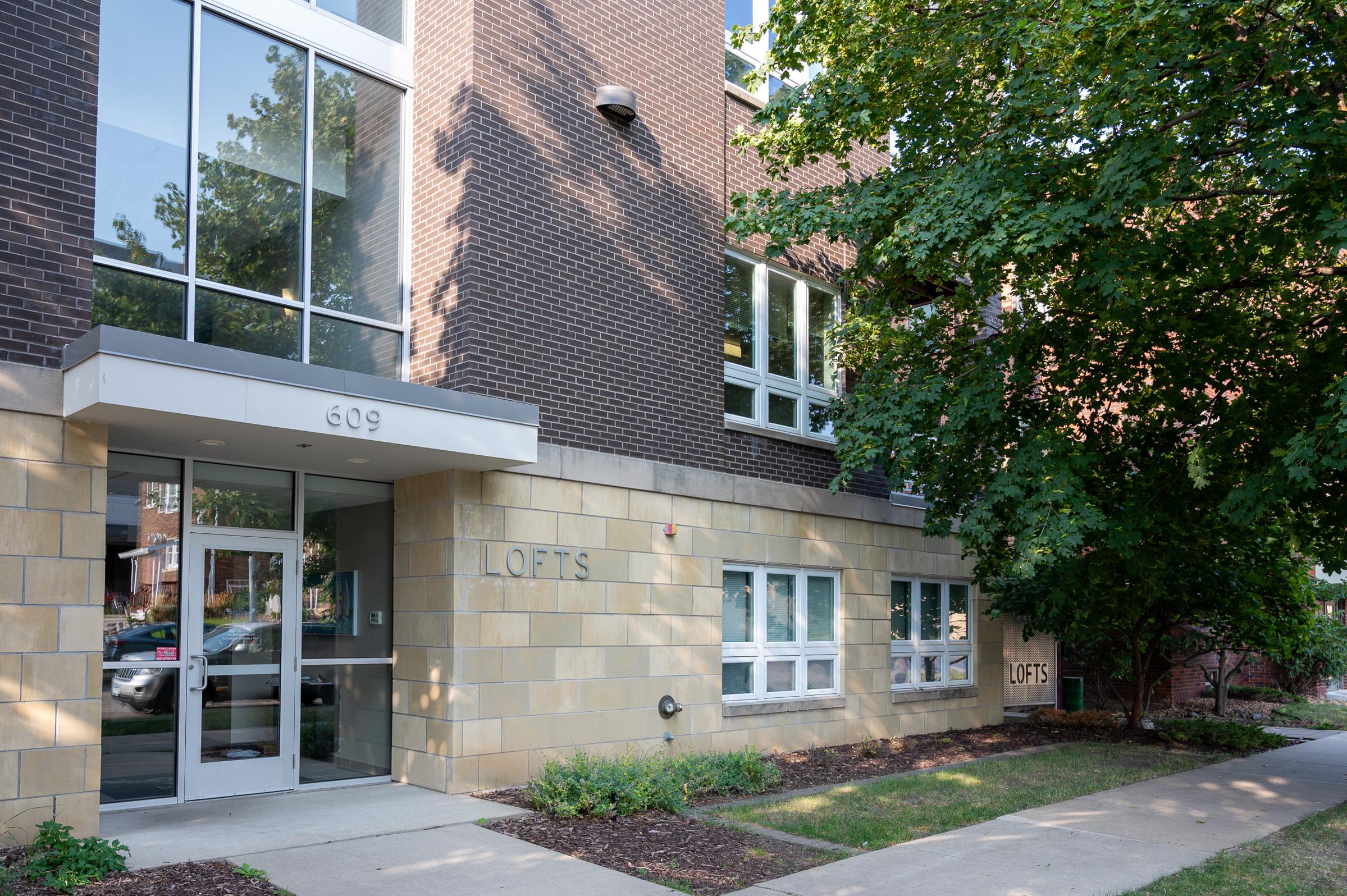 Exterior view of a modern brick building labeled "LOFTS." The building features large windows and a glass entrance. Surrounding the building are landscaped areas with trees and plants. The address "609" is visible above the entrance.