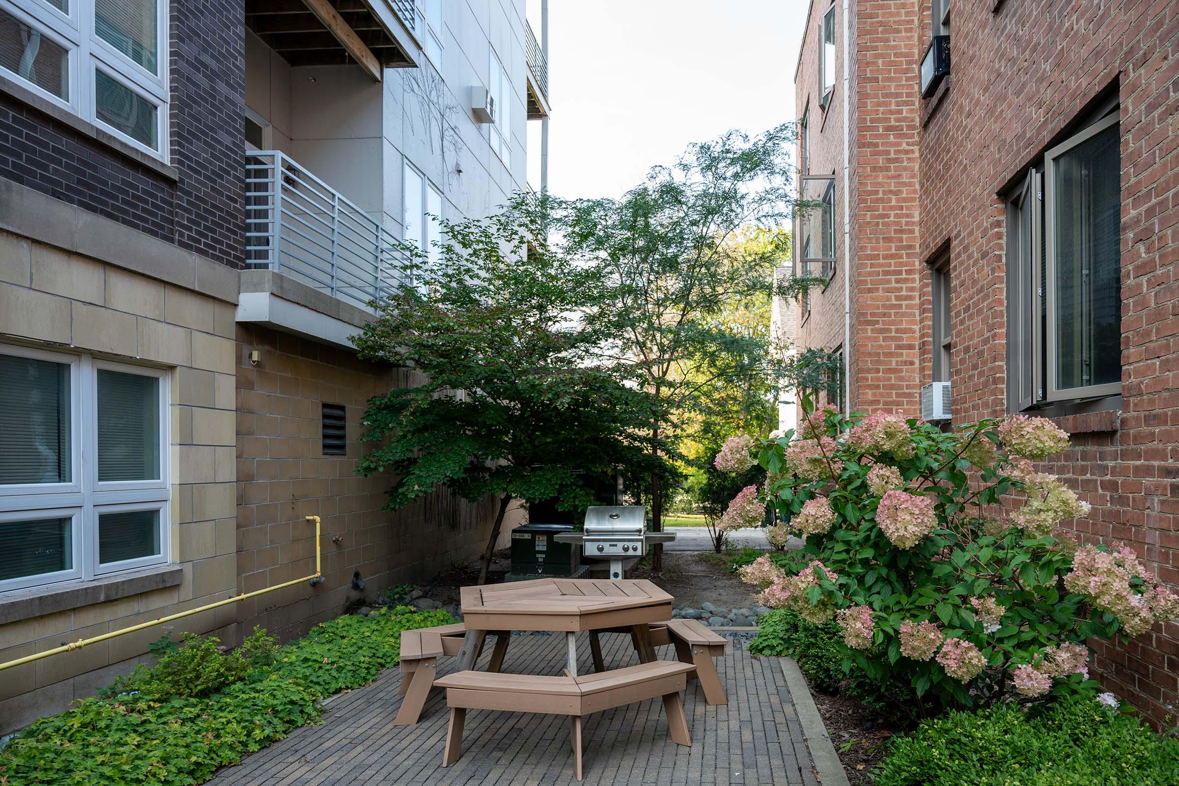 A narrow alleyway between two buildings featuring a wooden picnic table with matching benches, surrounded by greenery. A flowering bush with pink blooms is in the foreground, and a barbecue grill is visible in the background, creating a cozy outdoor space.