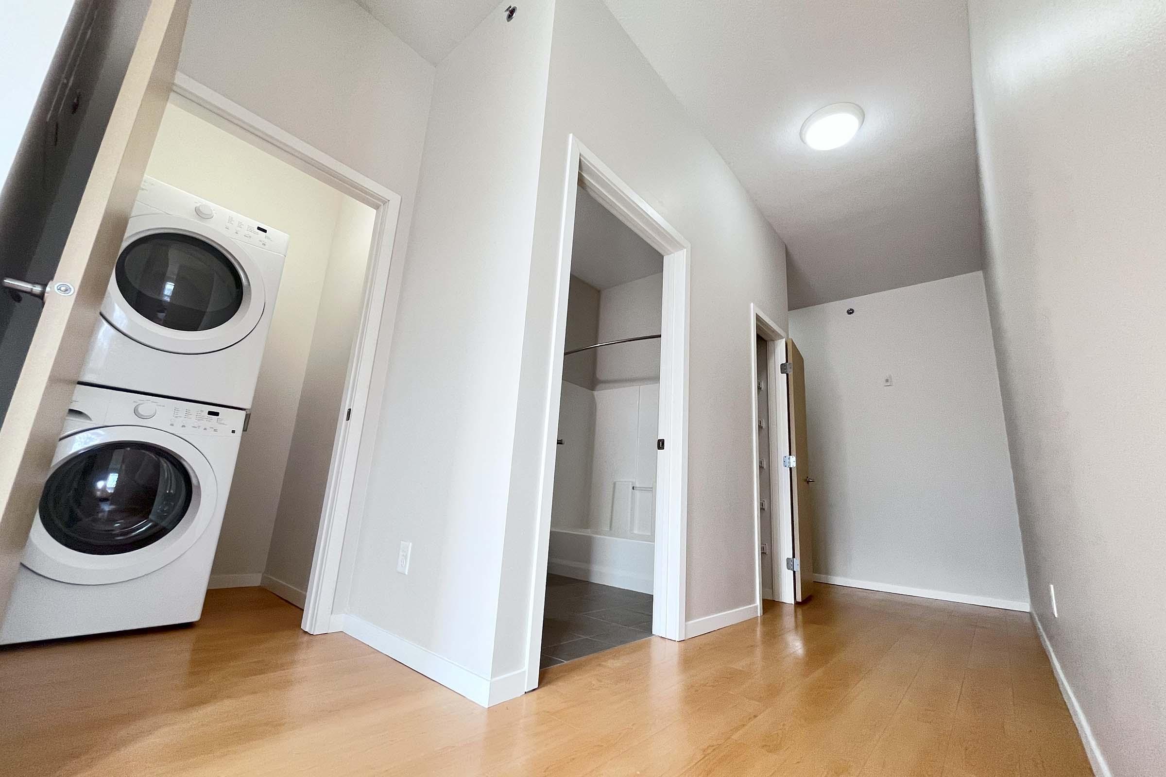 A bright and airy interior view of a laundry area featuring stacked white washing machines. A hallway leads to different rooms, with light-colored walls and wooden flooring. The space appears clean and modern, with doors on either side and a slight incline visible in the hallway.