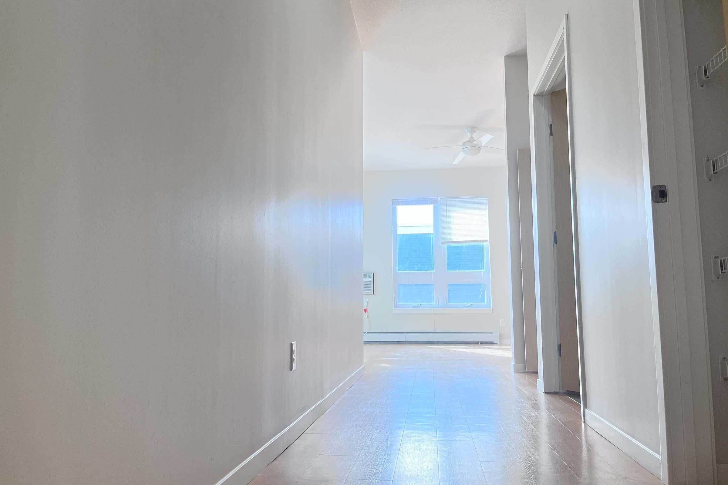 A light-filled hallway in a modern home with white walls and wooden flooring. The hallway leads to a bright room with large windows and a ceiling fan, providing a sense of openness and warmth. Doors to the left and right suggest access to additional rooms.