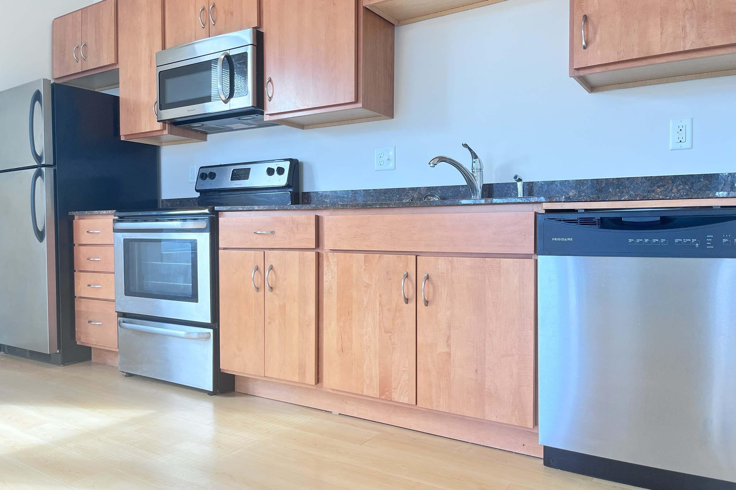 Modern kitchen featuring wood cabinets, a stainless steel microwave, an oven, and a stainless steel dishwasher. The countertop is dark, complementing the light-colored wooden floors and cabinetry. A silver refrigerator is visible on the left side.