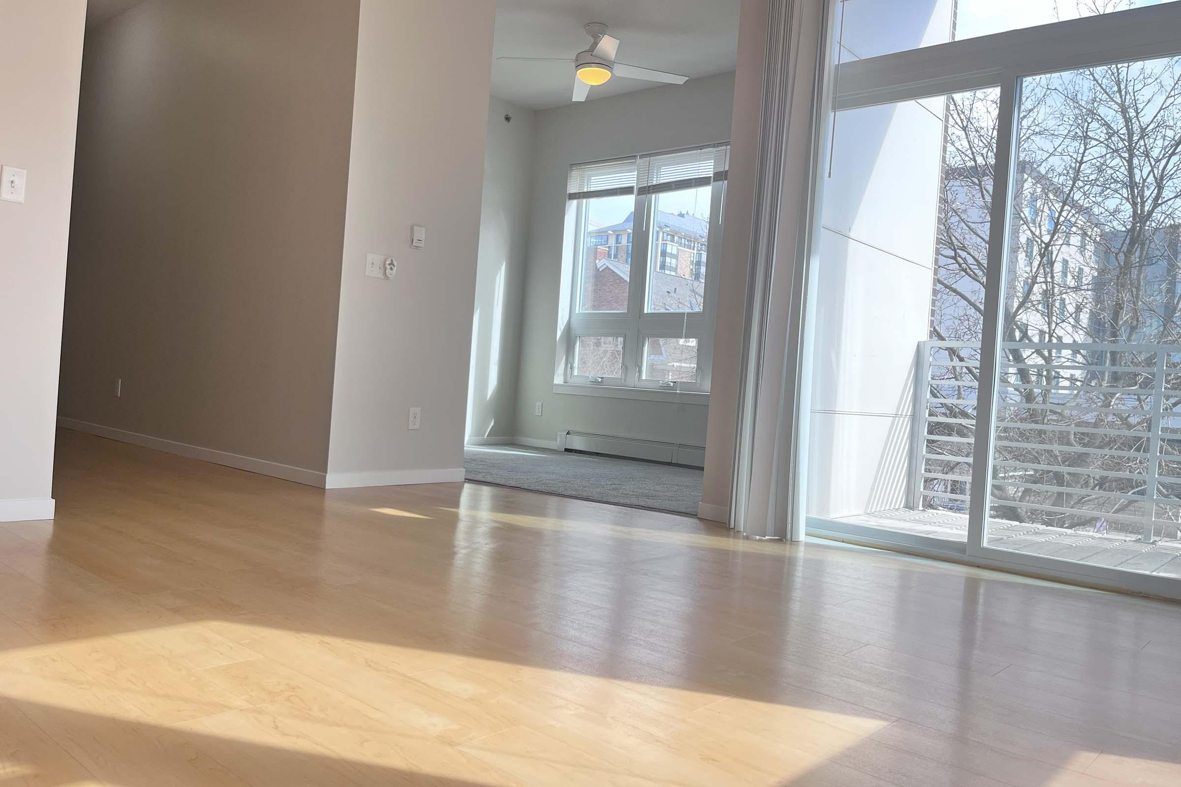 A spacious, modern interior view of a room with light-colored wooden flooring. Natural light streams in through large windows with white curtains. In the background, a balcony can be seen, and outside, trees and buildings are visible. The walls are painted a soft neutral color, creating a bright and inviting atmosphere.