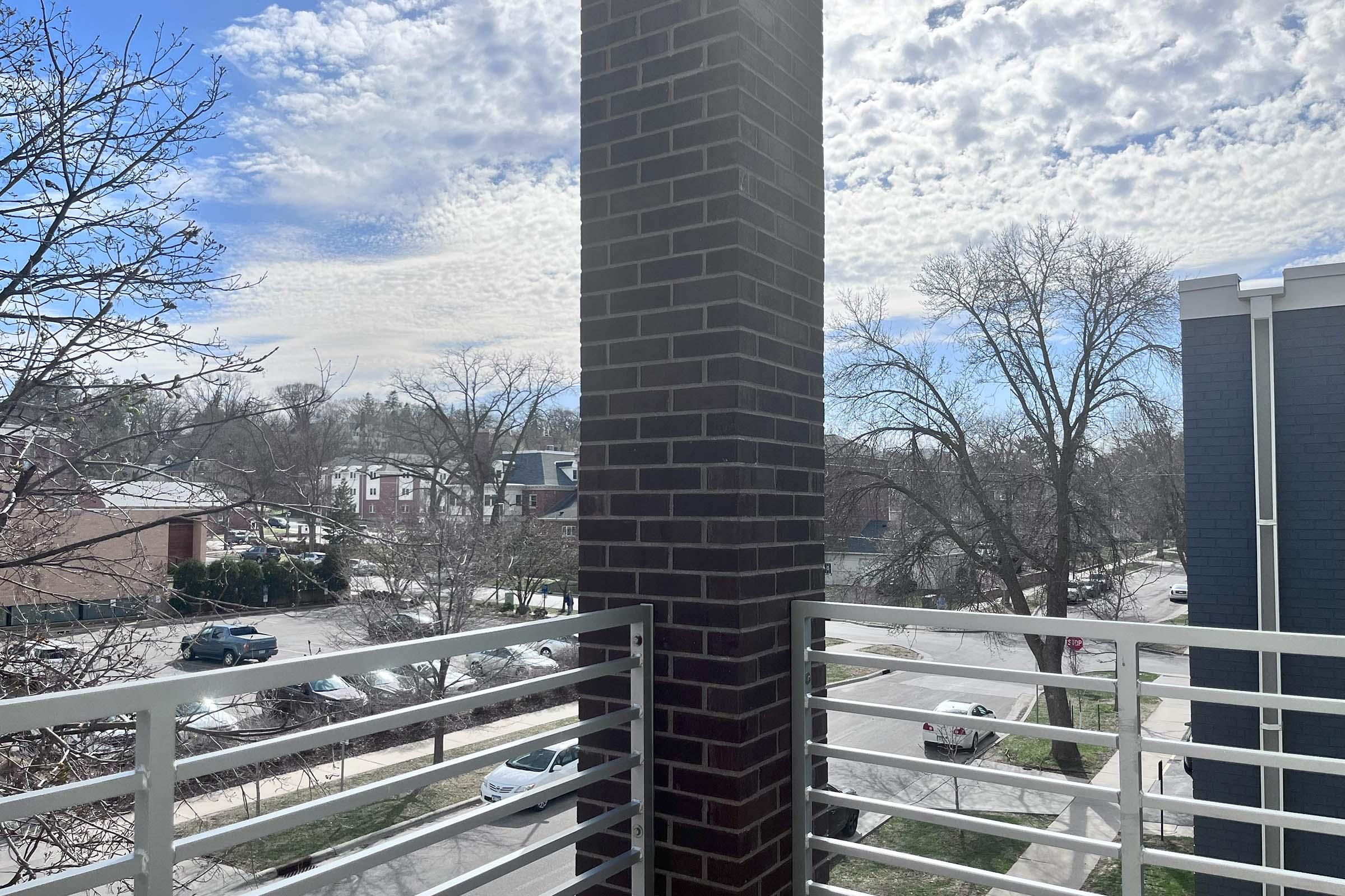 View from a balcony featuring a large brick column, with a blue sky filled with scattered clouds. In the background, there are trees and residential buildings, along with parked cars along the street, creating a serene urban landscape.