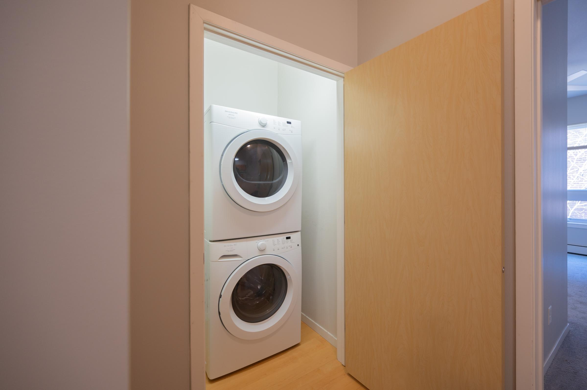 A laundry room featuring a stacked washer and dryer inside a small nook, with a light-colored door partially open, showing a minimalistic interior and wooden flooring. Natural light is indicated by a nearby window.