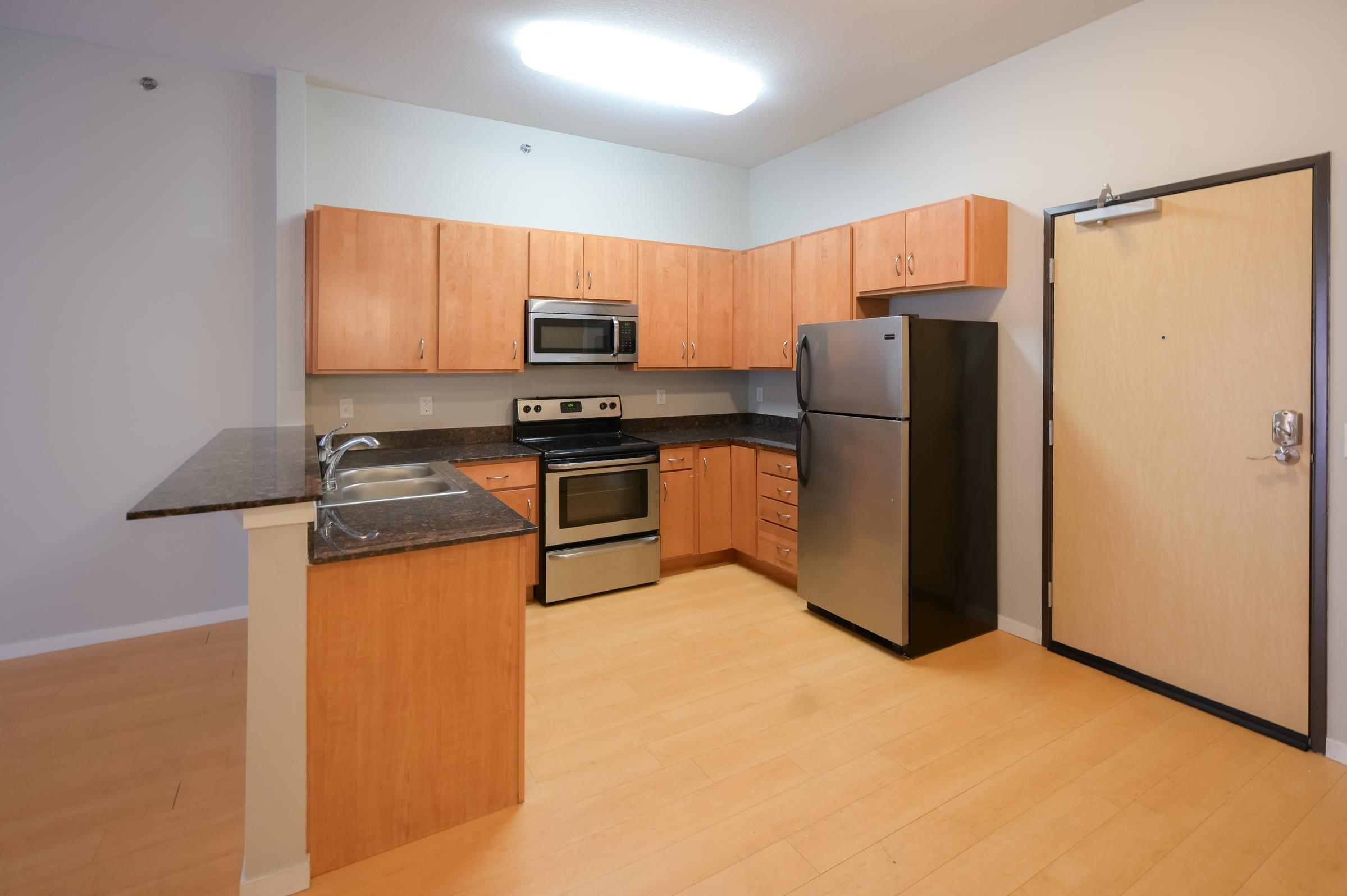 Modern kitchen with wooden cabinetry and stainless steel appliances, including a stove, refrigerator, and microwave. The countertop is dark granite, and the floor is light wood. There is an entrance door visible on the right side. Brightly lit with overhead lighting.