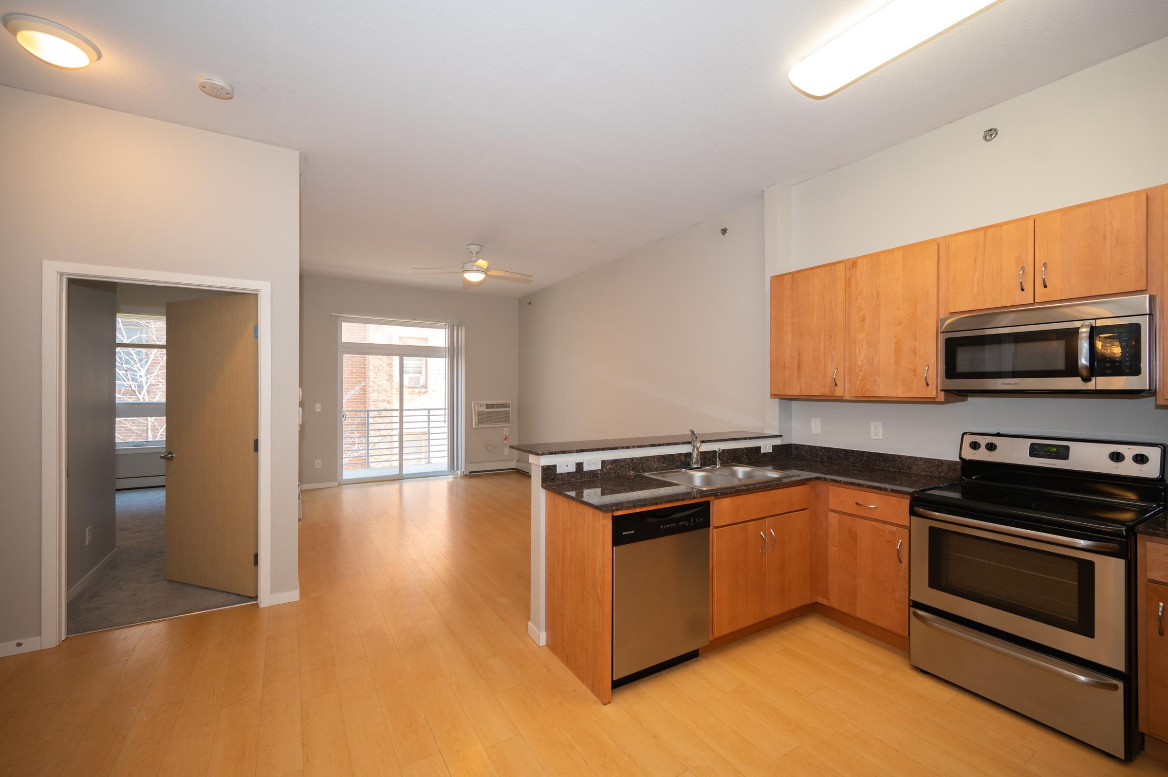 A modern kitchen featuring wooden cabinets, stainless steel appliances including a stove, oven, refrigerator, and dishwasher. The open layout leads to a living area with natural light. A doorway in the background provides access to a separate room, potentially a bedroom, with a balcony visible.