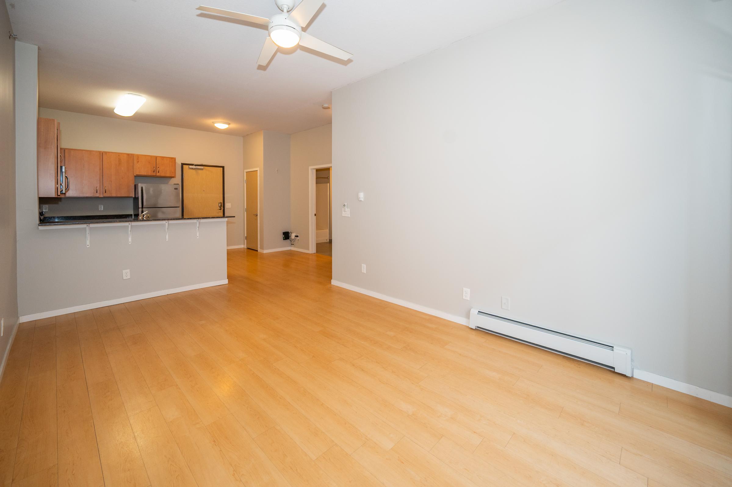 A spacious, empty room with light hardwood flooring, featuring a ceiling fan. In the background, there is an open kitchen with wooden cabinets and stainless steel appliances. The walls are painted a light gray, creating a bright and airy atmosphere. Doorways lead to other areas of the apartment.