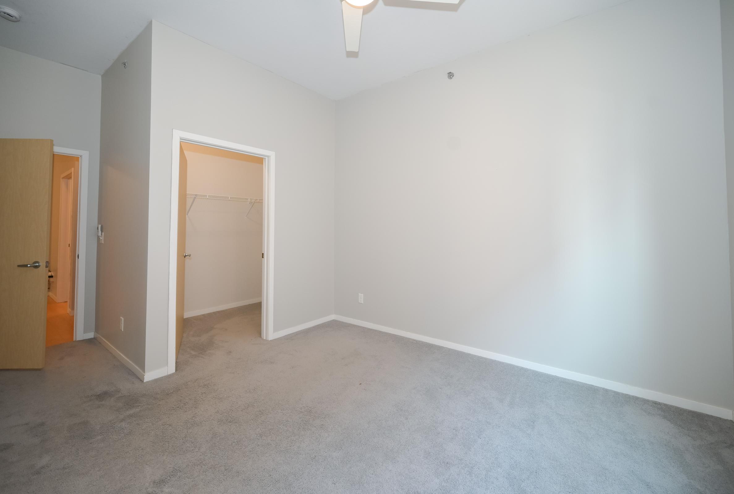 A spacious, empty room featuring light gray walls and carpet. On one side, there is a doorway leading to a closet with a hanging rack. A ceiling fan is visible above, and the room is well-lit with natural light coming from an unseen window. The overall ambiance is clean and minimalistic.