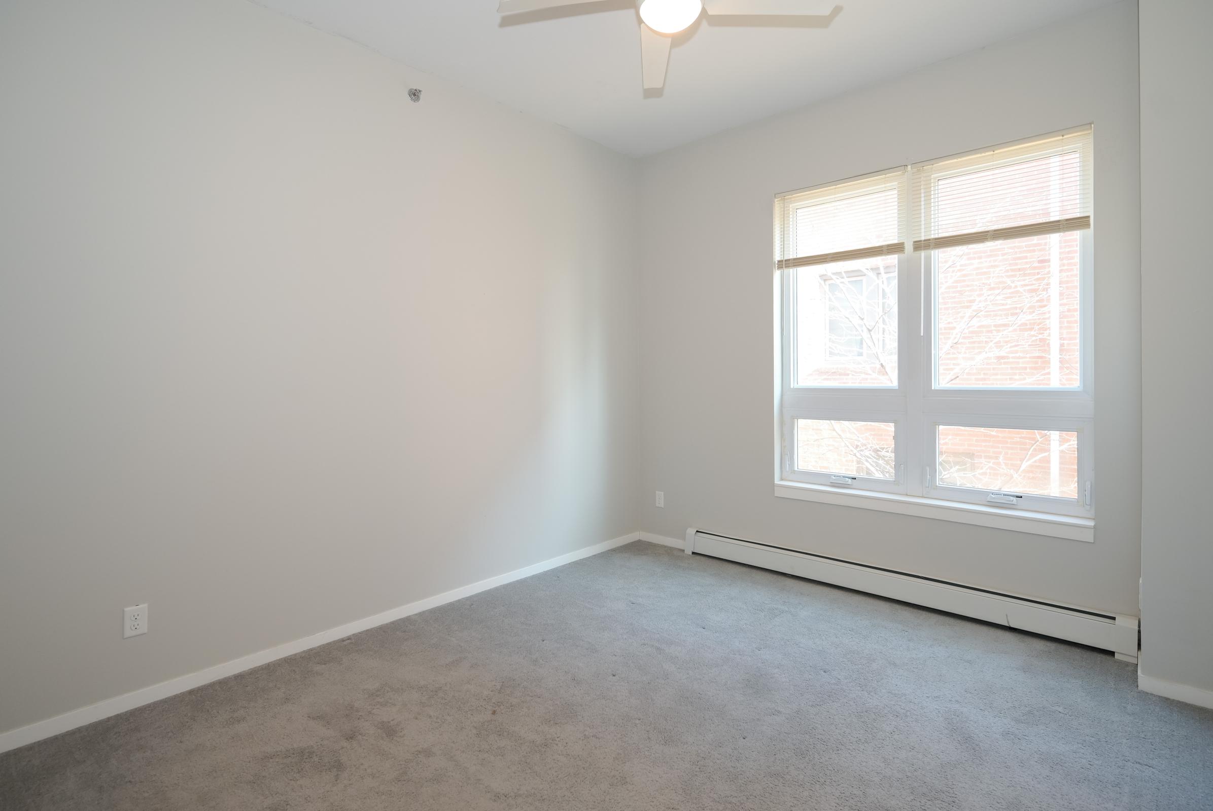 A spacious, empty room with light gray walls and a light carpet. The room features a ceiling fan and a large window with multiple panes, allowing natural light to fill the space. The room is unfurnished and has a clean, minimalist appearance.