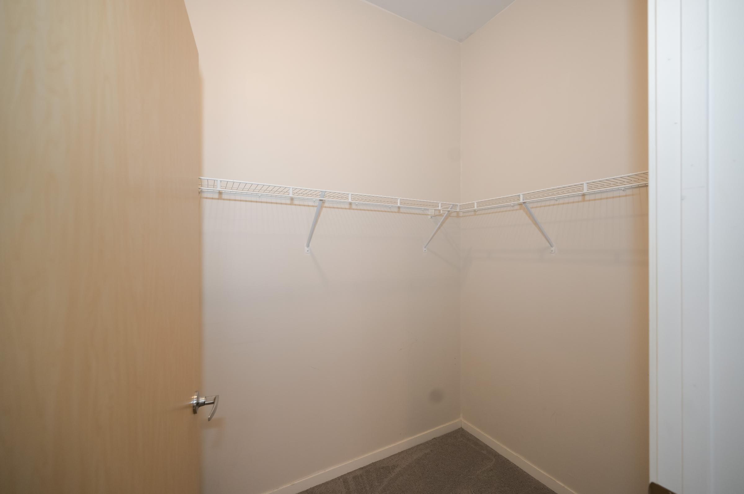 Empty closet space with beige walls and a gray carpet. One door is partially open, revealing a simple shelving system with two wire shelves for storage. The overall aesthetic is clean and minimalistic, focusing on the uncluttered interior.