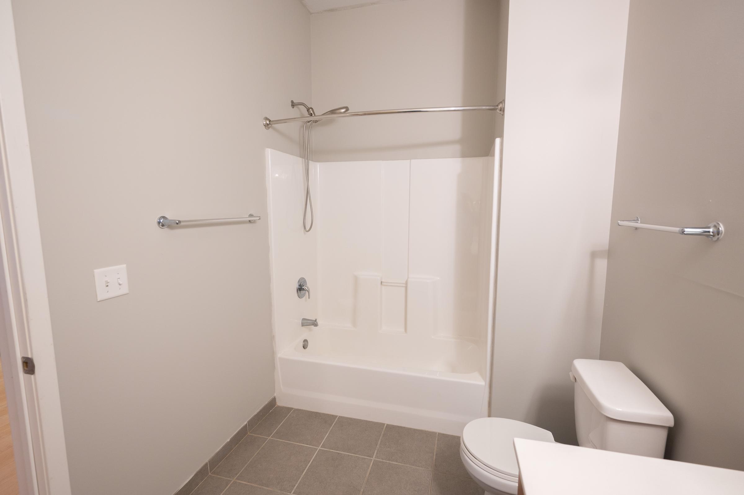 A clean, minimalist bathroom featuring a white bathtub with a showerhead, a toilet, and gray tile flooring. The walls are painted a light gray, with chrome towel bars mounted. The space is well-lit and uncluttered, creating a simple and modern look.
