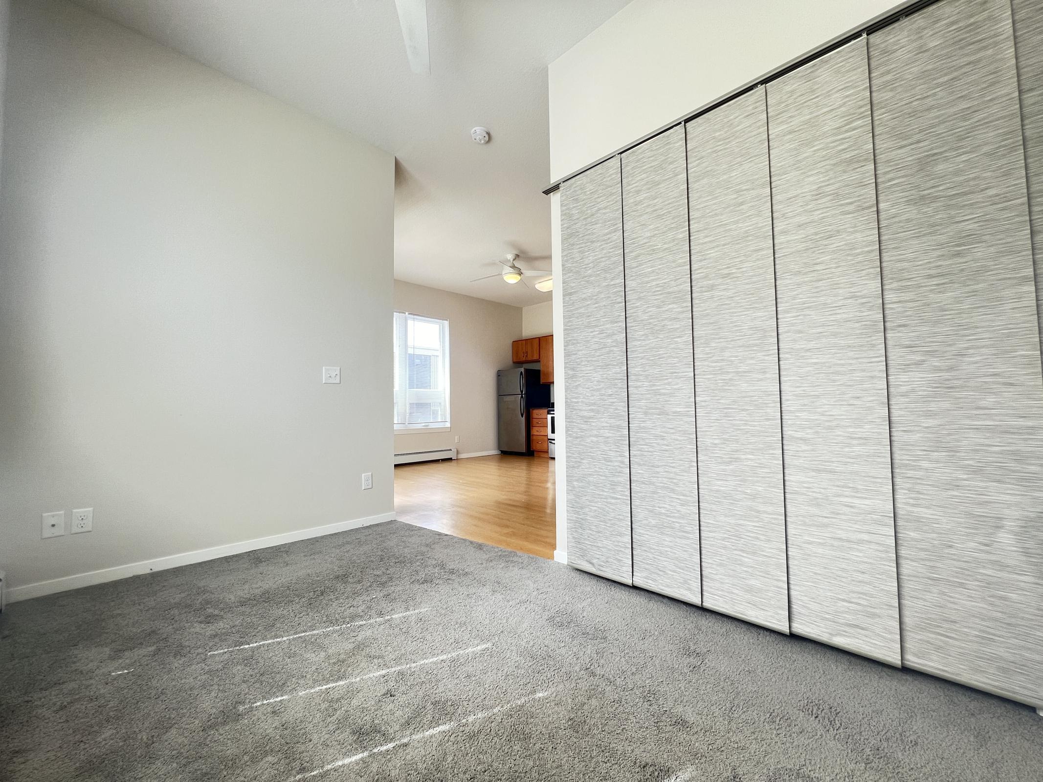Interior view of a room featuring gray carpet and a sliding closet door with a textured finish. In the background, there is a glimpse of a bright kitchen area with wooden cabinetry and ceiling fans, providing a spacious feel to the layout. Natural light is coming from a window.