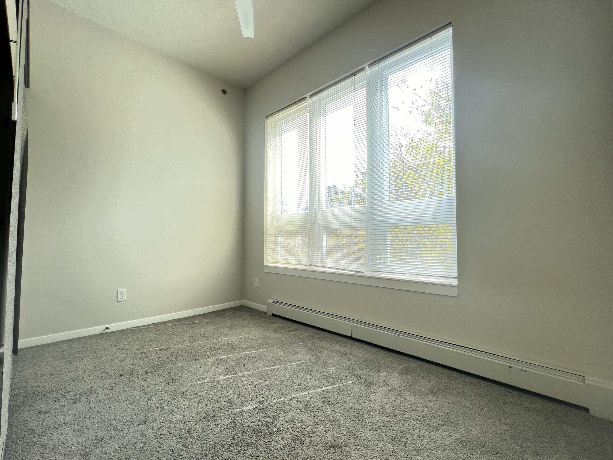 A bright, empty room with large windows allowing natural light to flow in. The walls are light-colored, and the carpet is gray. A ceiling fan is visible, and the floor has visible lines, possibly from furniture. The overall atmosphere is clean and minimalistic.