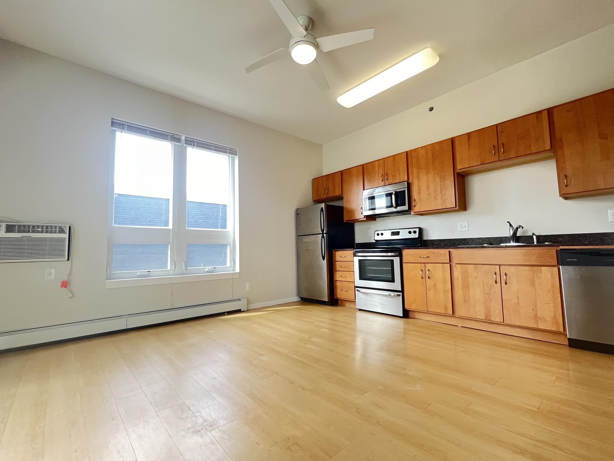 A modern kitchen with wooden cabinetry, stainless steel appliances including a refrigerator, stove, and microwave. There is a large window providing natural light, and a ceiling fan overhead. The floor is light-colored wood, and there is an air conditioning unit visible. Overall, the space appears bright and inviting.
