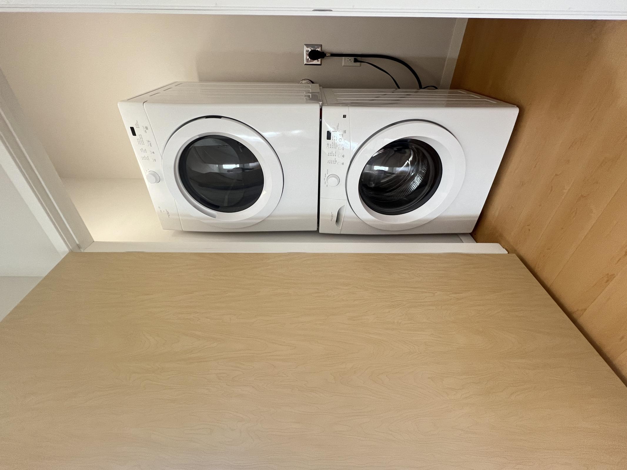 Stacked white washer and dryer in a small laundry space, with a light wooden door partially closed to the side. The appliances feature round glass doors and control panels at the top, set against a neutral-colored wall and wooden floor.
