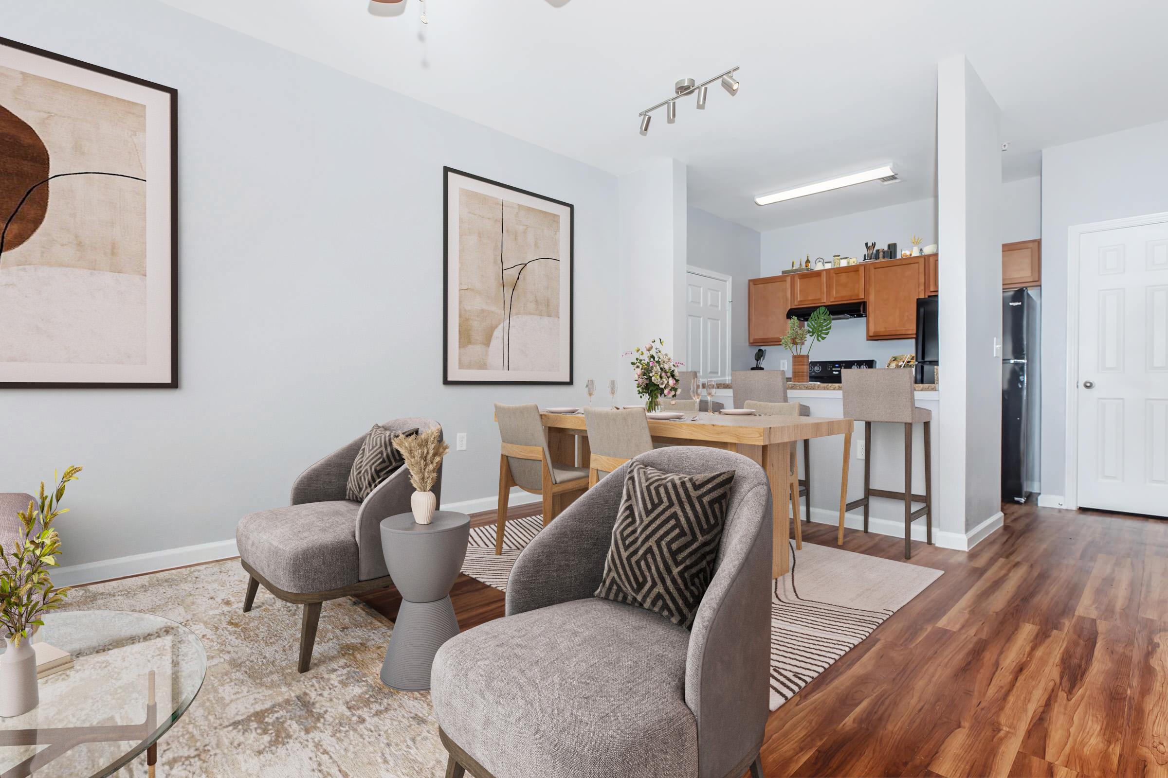 Modern living space featuring a light gray color scheme, two stylish chairs with geometric patterns, a wooden dining table with chairs, and abstract art on the walls. The kitchen is visible in the background, showcasing wood cabinetry and appliances, complemented by warm-toned hardwood flooring.