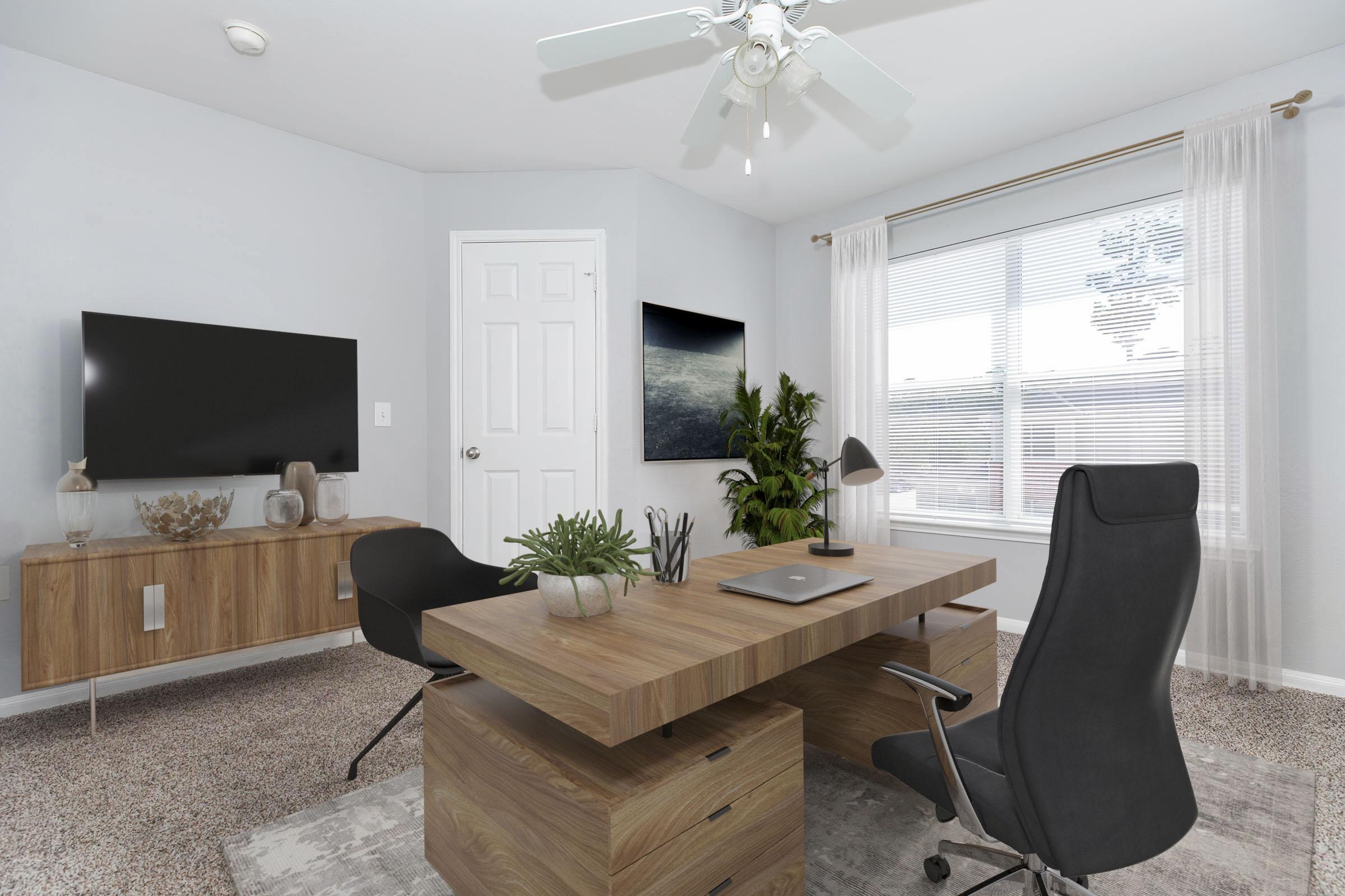 A modern home office featuring a wooden desk with a laptop, a black ergonomic chair, and a contemporary TV on the wall. There’s a potted plant, a decorative vase, and a large window with sheer curtains allowing natural light. The room has a neutral color scheme with carpet flooring.