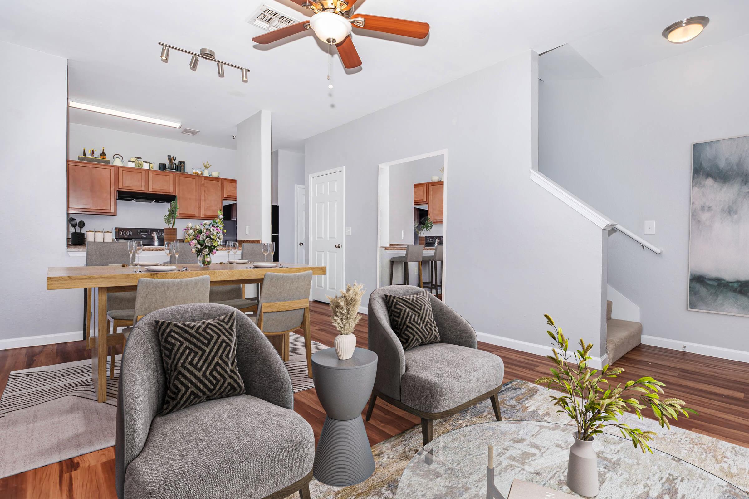 A modern living space with a dining area featuring a wooden table and gray chairs, stylish decor elements, and a staircase in the background. The room has light walls, wooden floors, and natural light, creating a welcoming atmosphere. Plants and decorative items enhance the aesthetics.