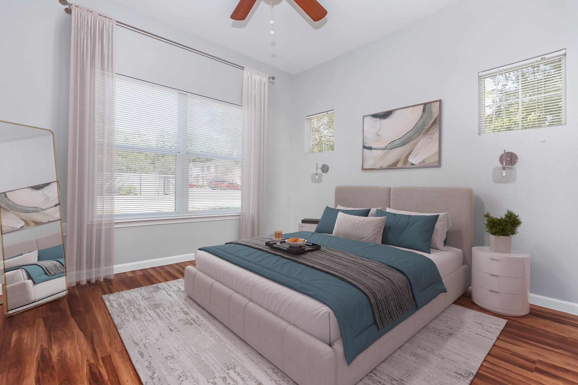 Cozy bedroom featuring a plush bed with teal and gray bedding, a stylish nightstand, and a modern mirror. Natural light streams in through large windows with blinds, illuminating the light-colored walls and hardwood floor, complemented by minimalistic artwork and decorative plants.