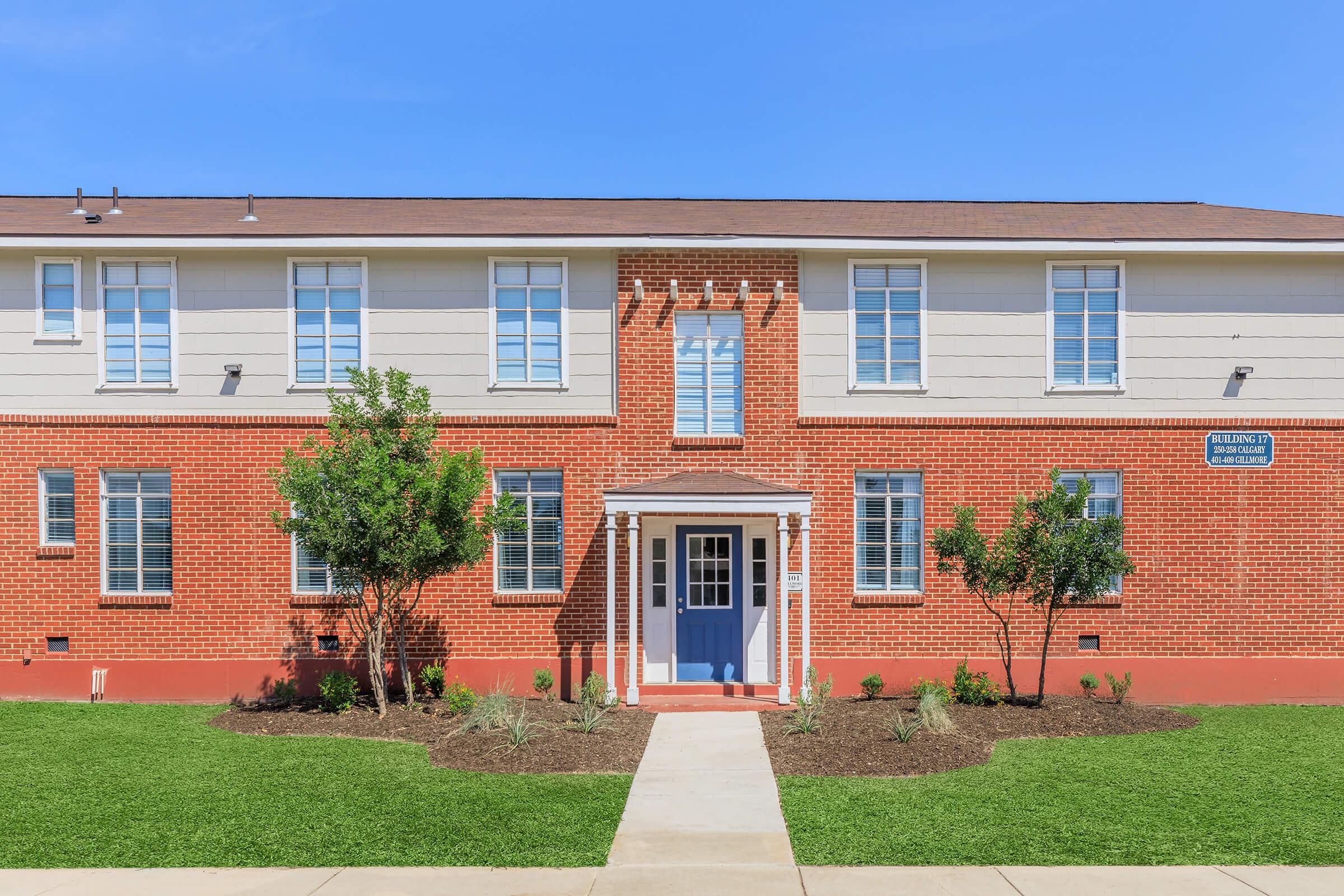 A two-story brick building with beige accents, featuring multiple windows and a centrally located blue door under a small porch. The surrounding area has neatly trimmed grass and landscaped bushes, with a clear blue sky in the background.