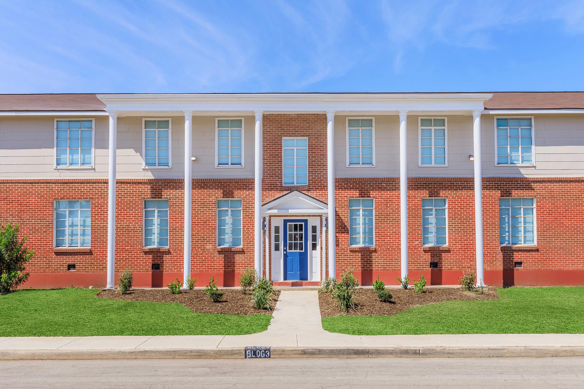 A modern two-story building with a red brick facade and white trim. The entrance features a blue door and is flanked by symmetrical windows. The landscape includes green grass and small plants, creating a welcoming outdoor space under a clear blue sky.