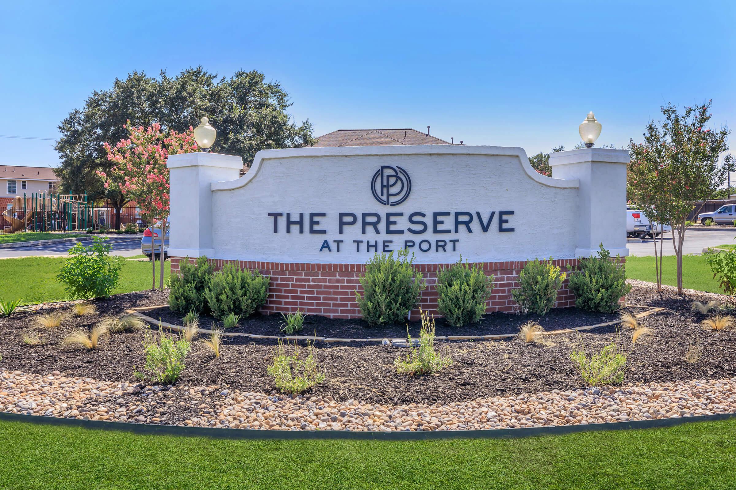 Sign for "The Preserve at the Port" featuring a white stucco and brick design. The sign is surrounded by landscaped greenery, including shrubs and small plants, with a clear blue sky in the background.