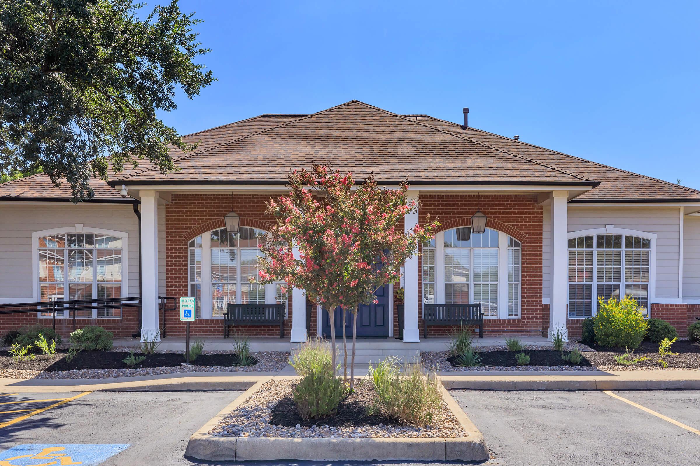 A well-maintained building with a peaked roof and brick facade. It features large windows on either side of the entrance, framed by tall columns. In front, there is a flowering tree and landscaped shrubbery, with parking spaces visible in the foreground under a clear blue sky.