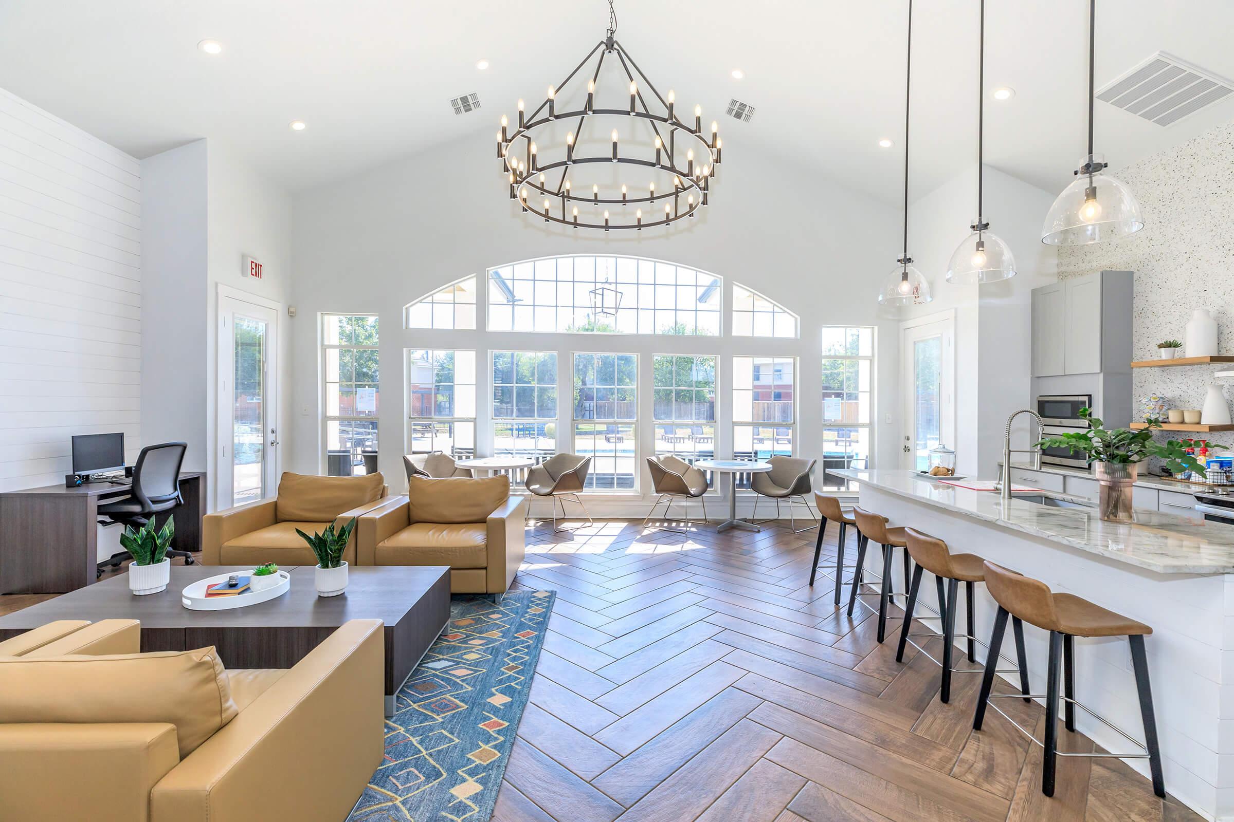 A modern, airy lounge area featuring comfortable brown leather sofas, a coffee table, and a decorative rug. Large windows provide ample natural light, with a view of an outdoor space. A kitchen bar with seating, elegant light fixtures, and a small workspace complete the inviting atmosphere.