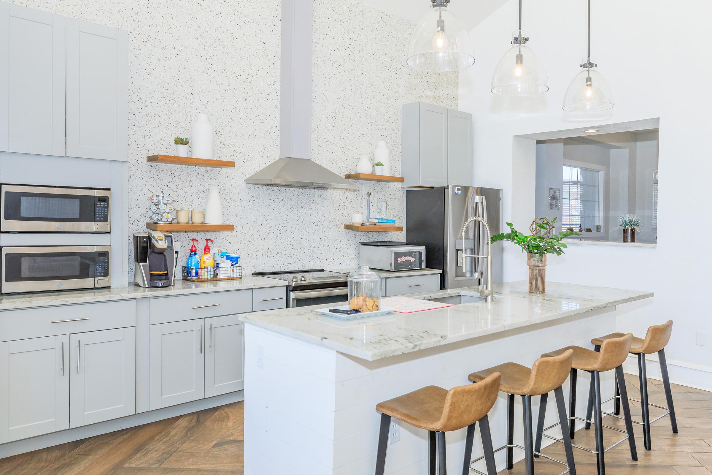 Modern kitchen featuring a large island with four wooden bar stools, gray cabinets, stainless steel appliances, and a speckled wall. Natural light illuminates the space, highlighting decor elements like plants and kitchen essentials on the countertop and shelves.