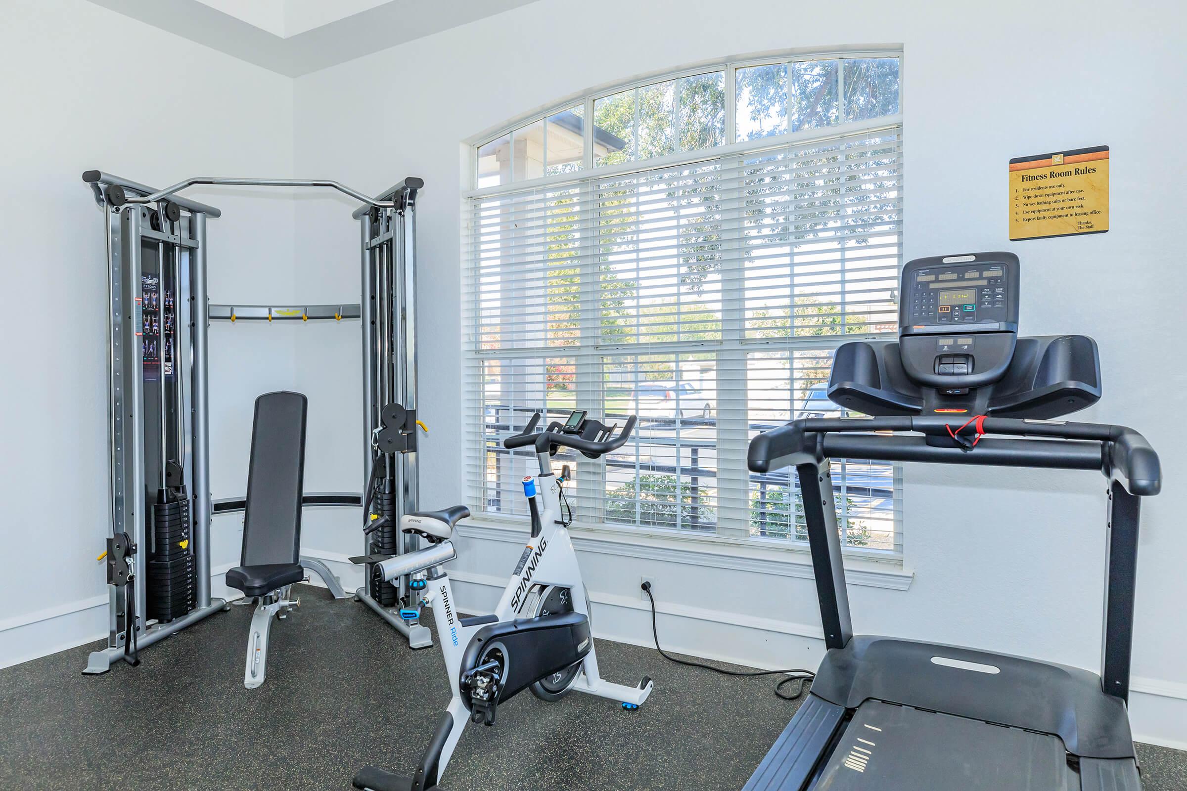 A bright fitness room featuring a multi-station weight machine, a stationary bike, and a treadmill. Large windows allow natural light to flood the space, with trees visible outside. The walls are painted in light colors, creating an inviting atmosphere for workouts.