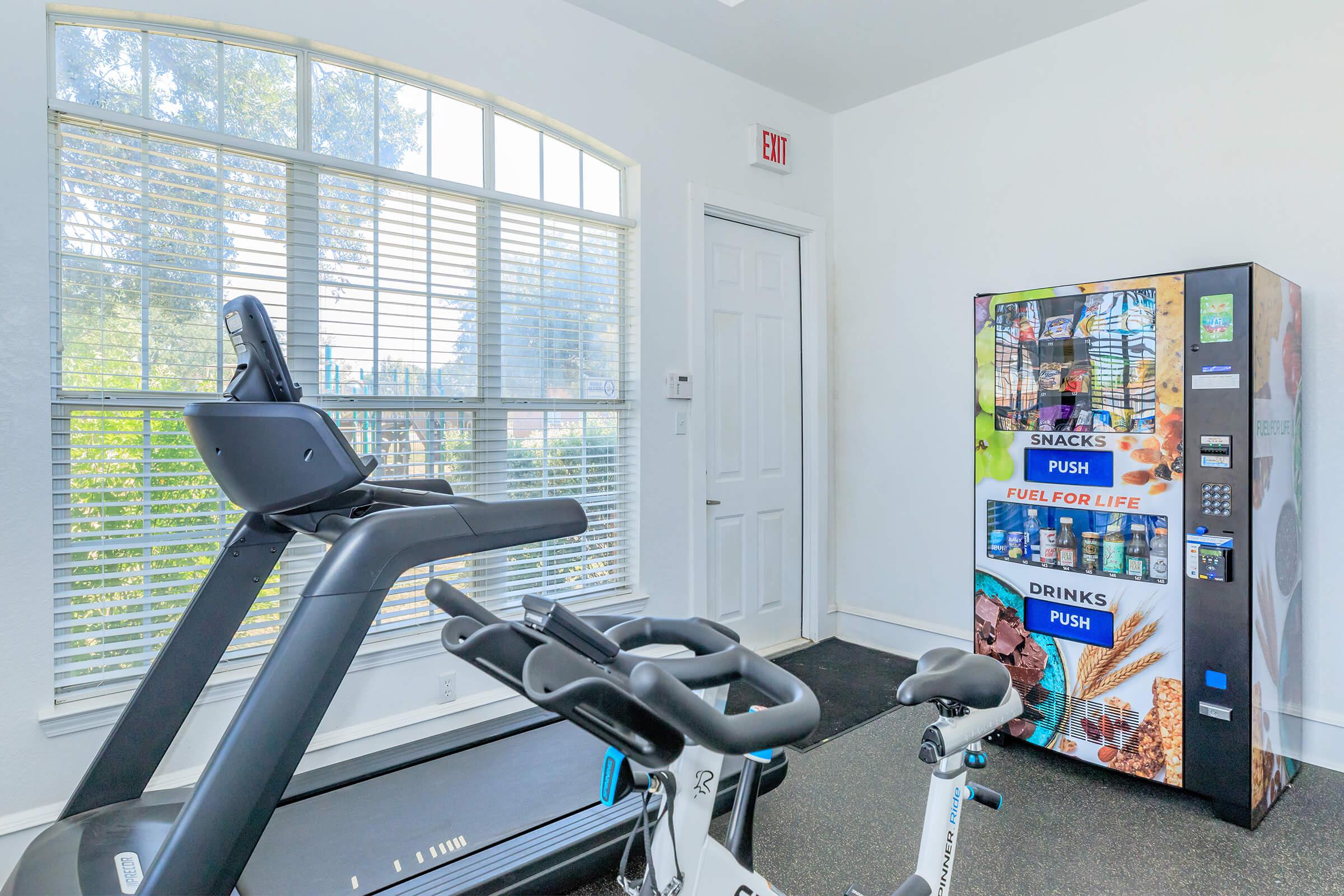 A fitness room featuring a treadmill and stationary bike. There is a vending machine displaying snacks and drinks nearby, with large windows allowing natural light to brighten the space. The walls are painted light gray, creating a modern and inviting atmosphere for workouts.