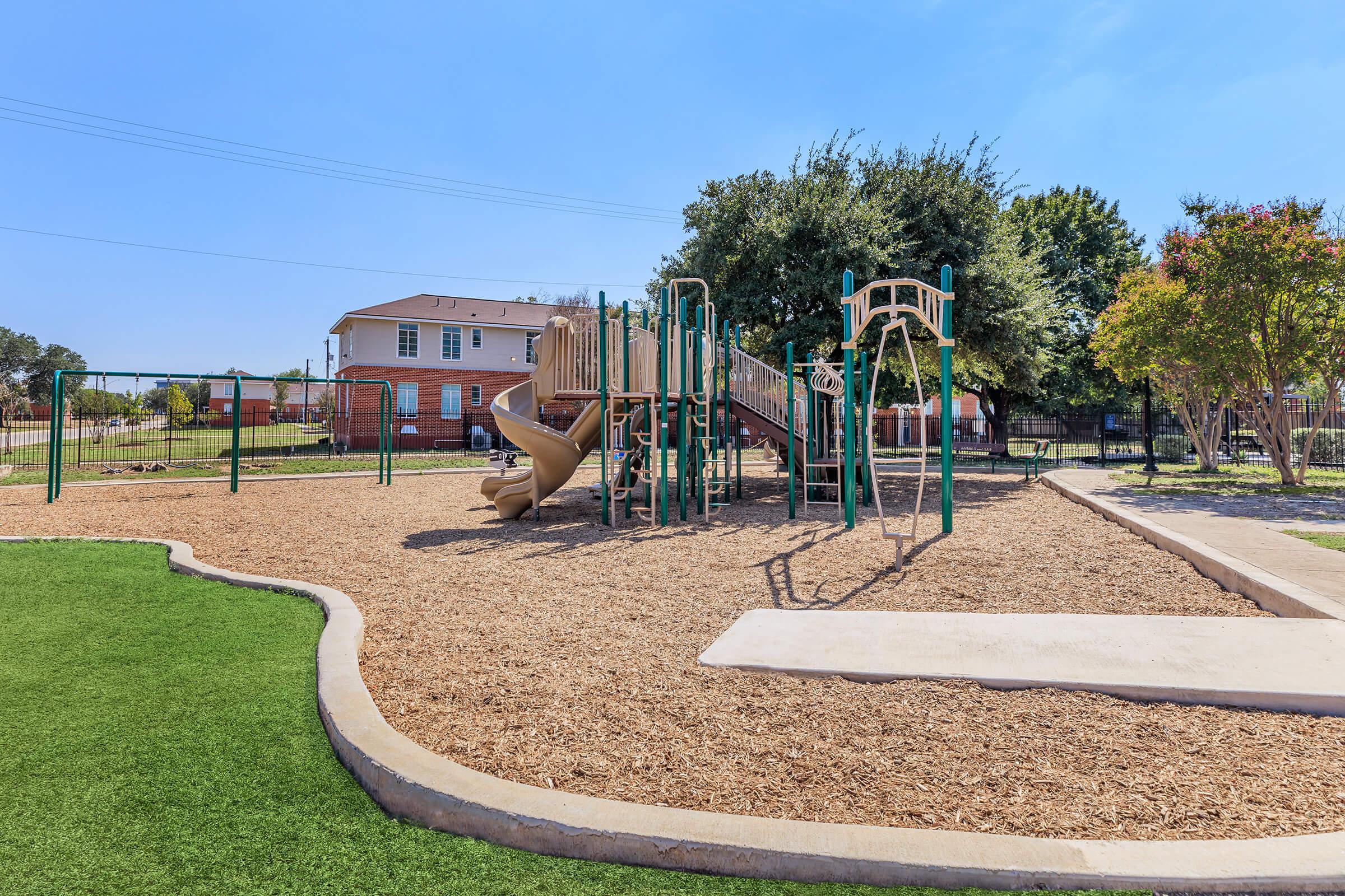 A playground area featuring a wooden climbing structure with a slide, swings, and safety mulch flooring. Surrounded by green grass and trees, with residential buildings in the background under a clear blue sky.