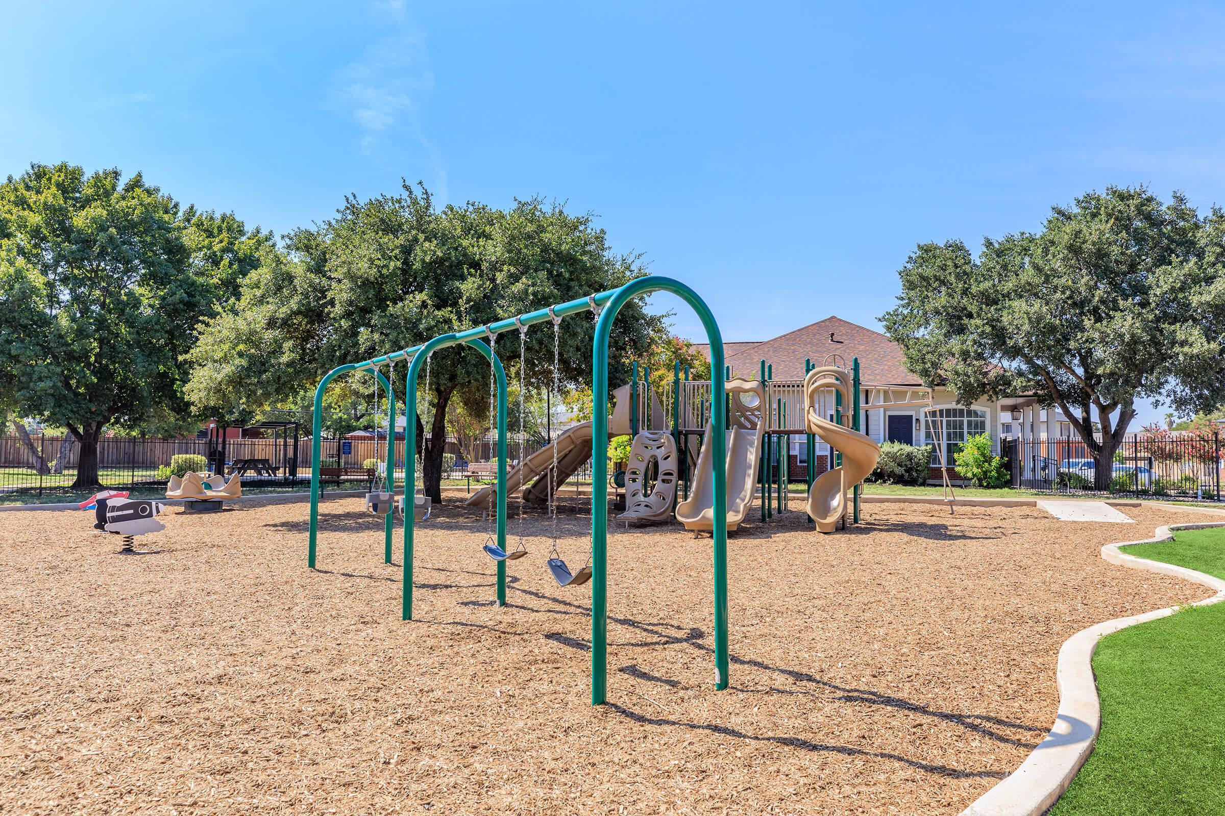 A bright, sunny playground featuring swings, a slide structure, and a soft, mulch-covered ground. In the background, there are trees providing shade and a building that appears to be a community center or residential area. The sky is clear with a few clouds.